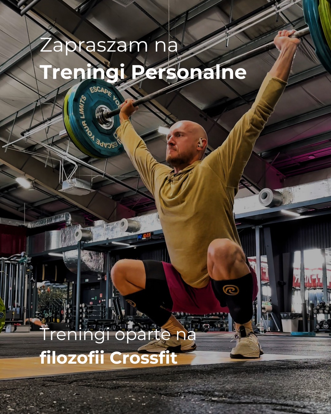 Mężczyzna w przysiadzie z uniesioną sztangą nad głową w siłowni, prezentujący trening personalny oparty na filozofii Crossfit. Widoczne obciążenia i wyposażenie siłowni.