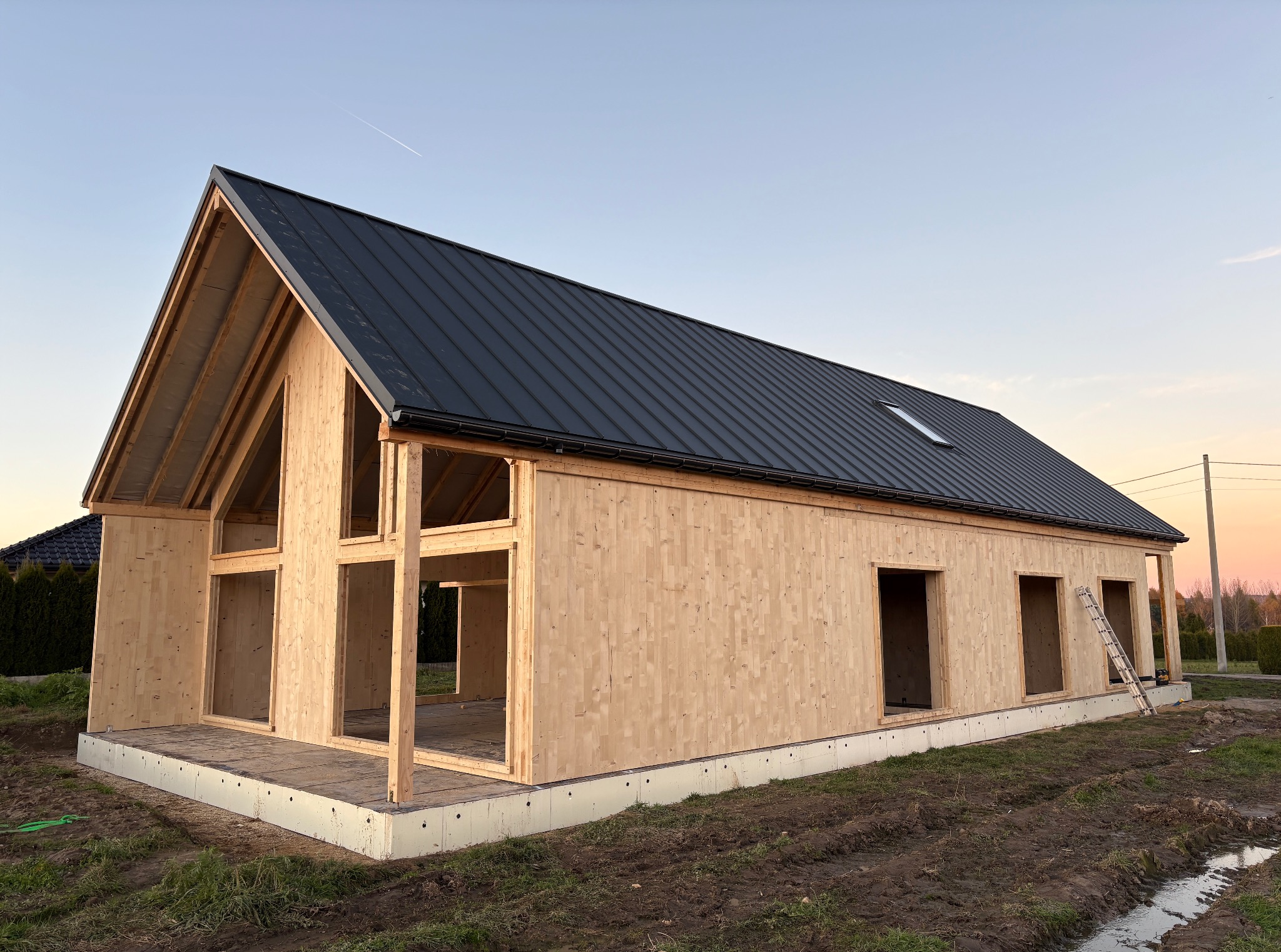 Surowy stan domu z drewna klejonego warstwowo, z czarnym dachem z blachy na tle błękitnego nieba o zmierzchu. Widoczne drewniane ściany i konstrukcja dachu.