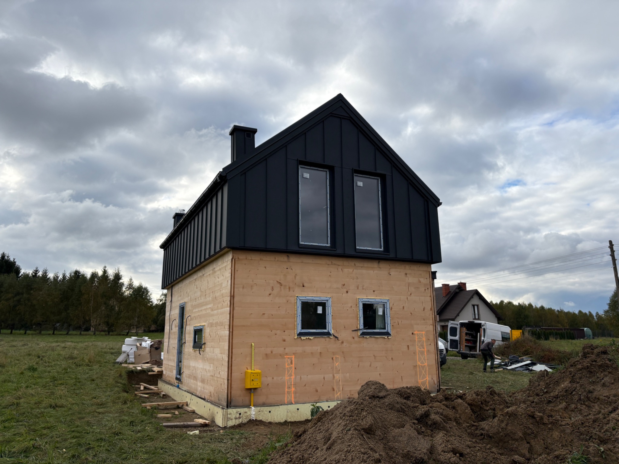 Surowy dom z czarnym dachem blisko ukończenia, z widoczną drewnianą elewacją i przygotowanym terenem. W tle pochmurne niebo i częściowo widoczny samochód dostawczy.