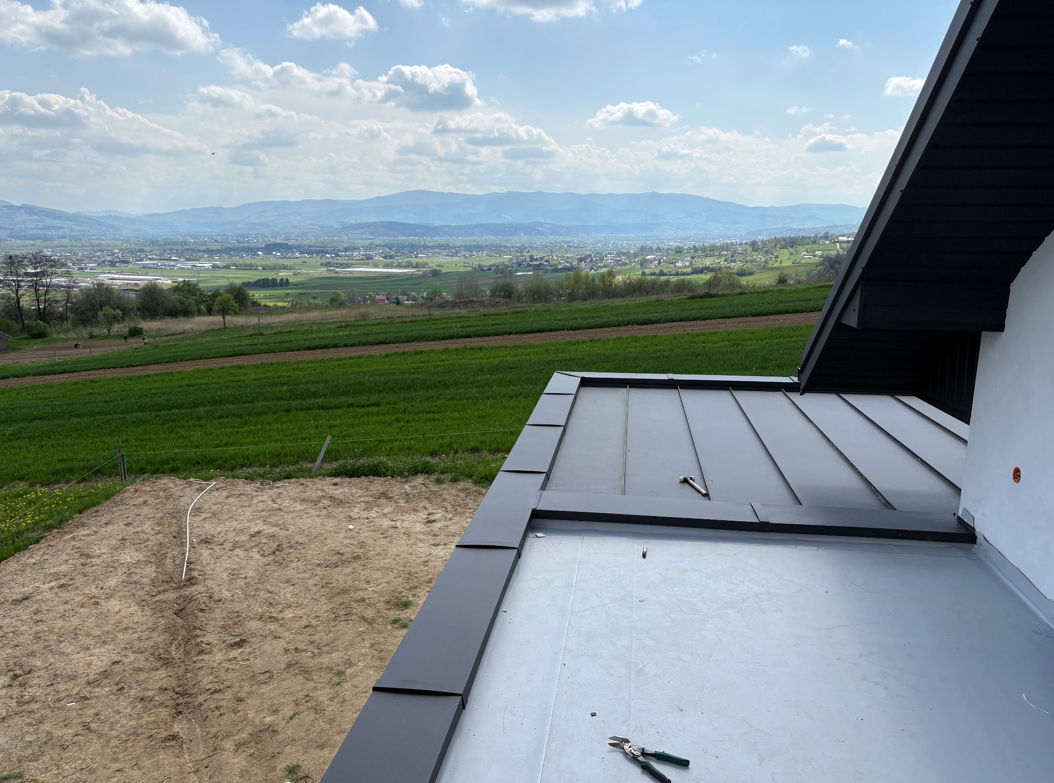 Nowoczesny dach z blachy na tle malowniczego krajobrazu. Widoczne narzędzia dekarskie, co sugeruje etap prac wykończeniowych. Dach w kolorze antracytowym.