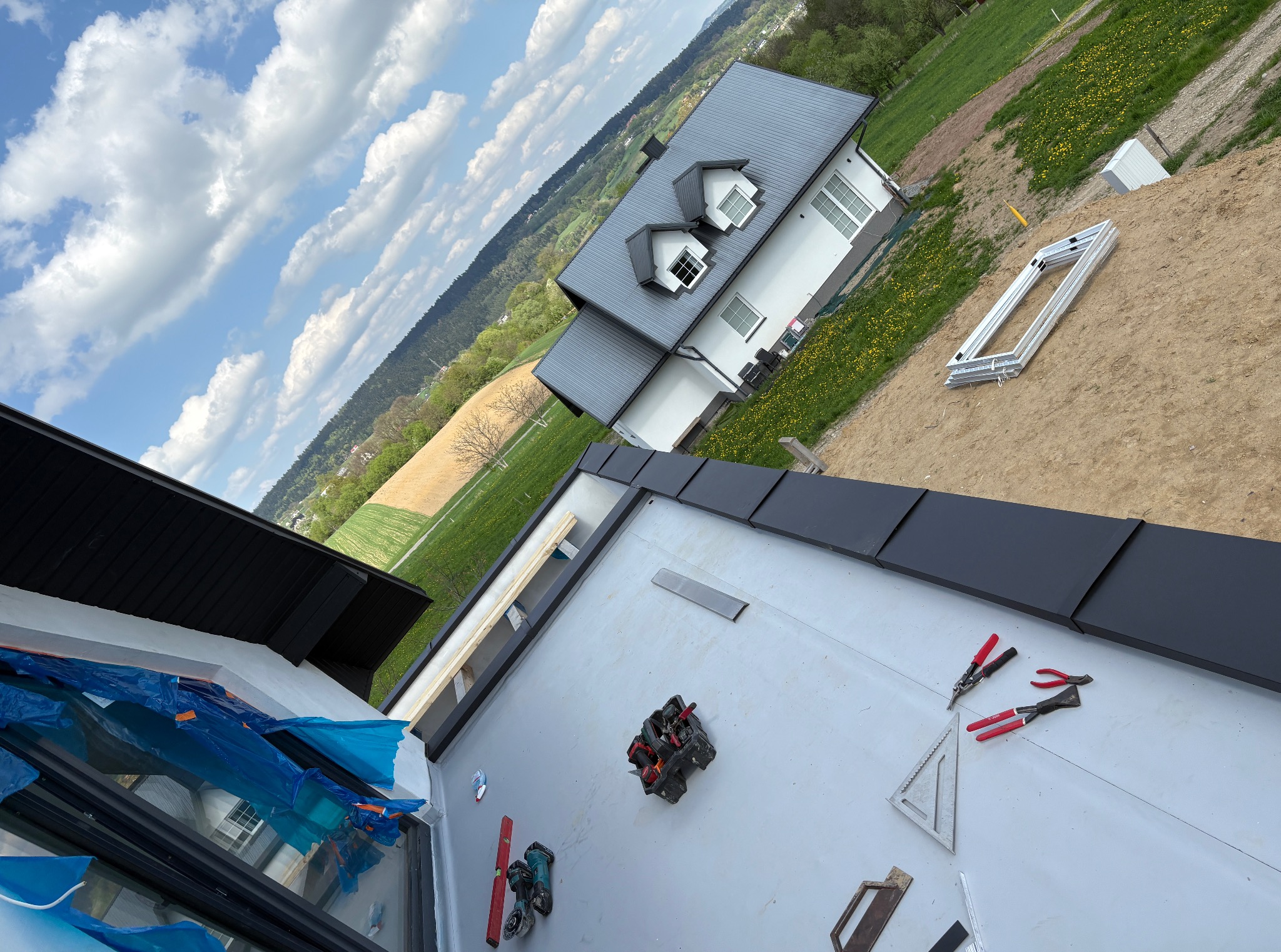 Montaż dachu z blachy na domu jednorodzinnym. Widoczne narzędzia dekarskie, fragmenty dachu i krajobraz w tle. Ujęcie z góry, ukośne.