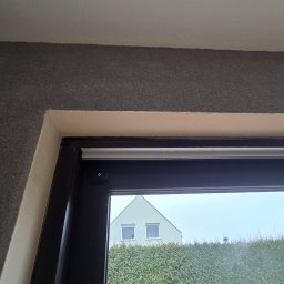 Portals - Dawid Bondos Okna Drzwi Bramy - Fragment wnętrza z oknem, widok na dom przez szybę. Ściana z szarą strukturą, białe wykończenie ramy okiennej. Widoczne detale montażu.