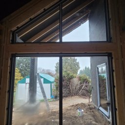 Portals - Dawid Bondos Okna Drzwi Bramy - Nowoczesne okno z czarną ramą, widok z wnętrza surowego budynku na zewnątrz. W tle zielony namiot i drzewa. Widoczne drewniane elementy konstrukcyjne.