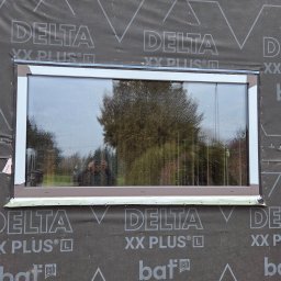 Portals - Dawid Bondos Okna Drzwi Bramy - Montaż okna z brązową ramą w nowym budynku, widoczne zabezpieczenie folią Delta XX Plus. W szybie odbija się osoba robiąca zdjęcie.