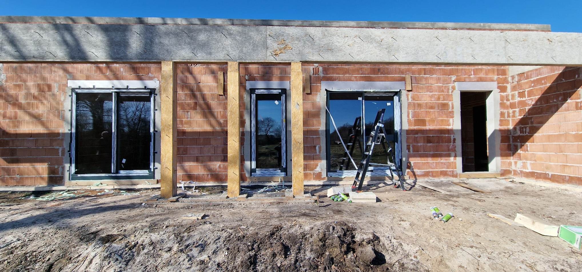 Budowa domu w trakcie montażu okien i drzwi. Widoczne okna z szybami refleksyjnymi, drabina i materiały budowlane. Surowy stan, cegła i ziemia.