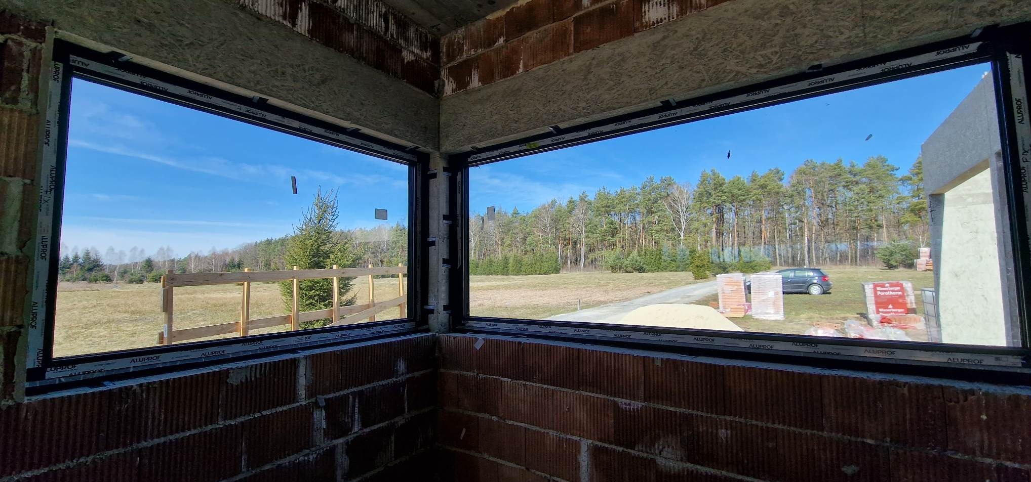 Dwa okna PCV z czarnymi ramami w surowym murze, widok na zewnątrz: las, łąka, sterta piasku i cegły Porotherm. Okna marki Aluprof.