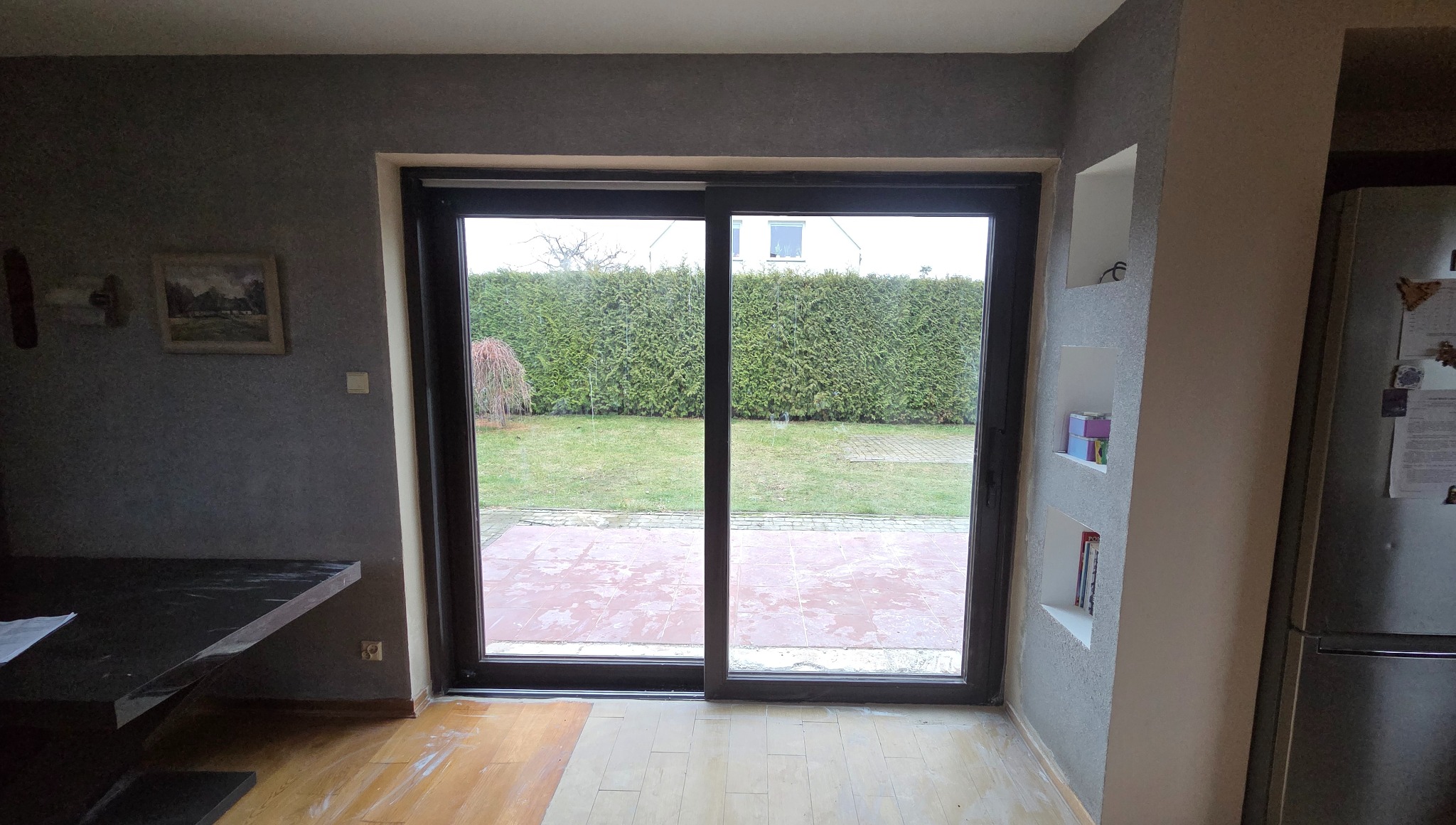 Ciemne, przesuwne drzwi tarasowe z widokiem na ogród, z niskim progiem. Wnętrze z szarą ścianą i drewnianą podłogą. Minimalistyczny design.