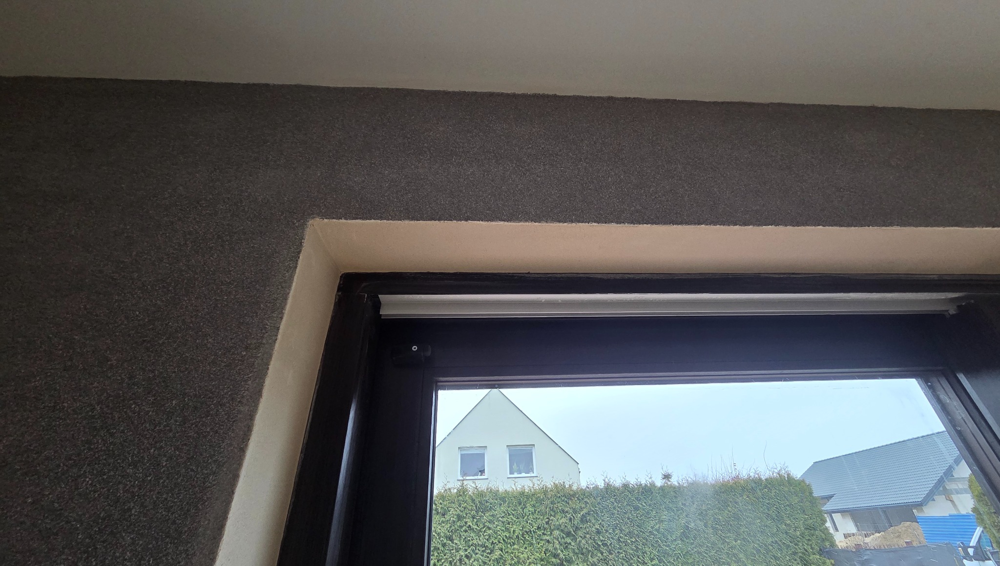 Fragment wnętrza z oknem, widok na dom przez szybę. Ściana z szarą strukturą, białe wykończenie ramy okiennej. Widoczne detale montażu.