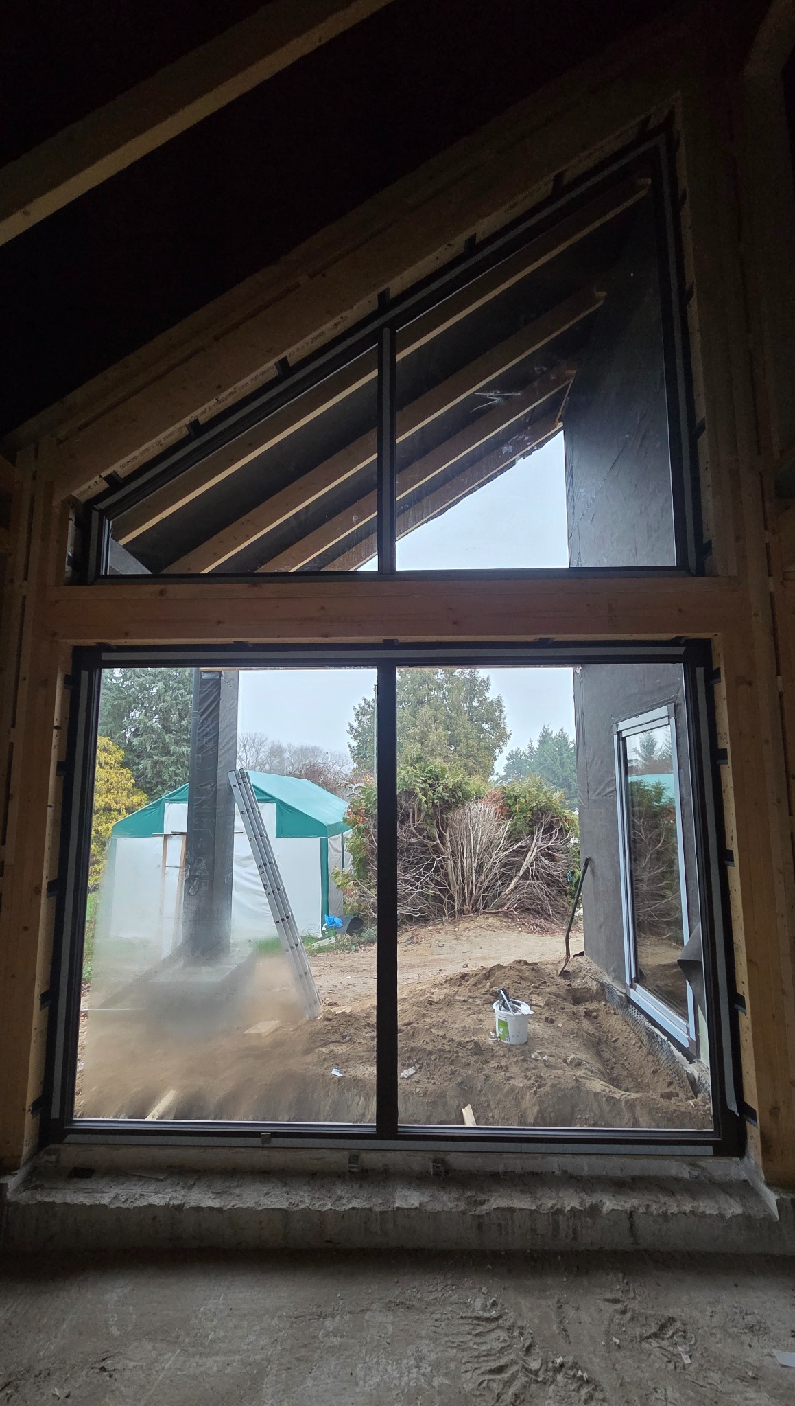 Nowoczesne okno z czarną ramą, widok z wnętrza surowego budynku na zewnątrz. W tle zielony namiot i drzewa. Widoczne drewniane elementy konstrukcyjne.