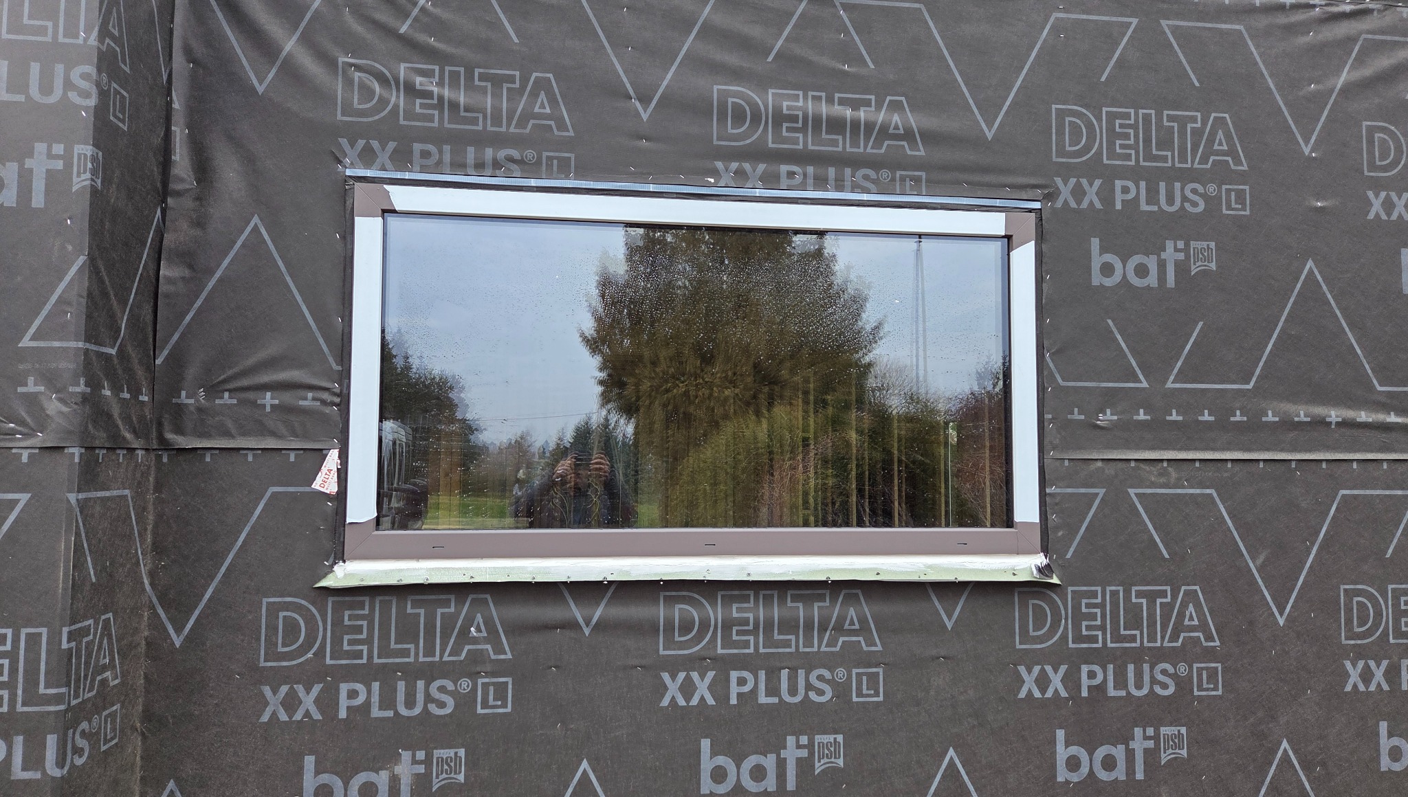 Montaż okna z brązową ramą w nowym budynku, widoczne zabezpieczenie folią Delta XX Plus. W szybie odbija się osoba robiąca zdjęcie.