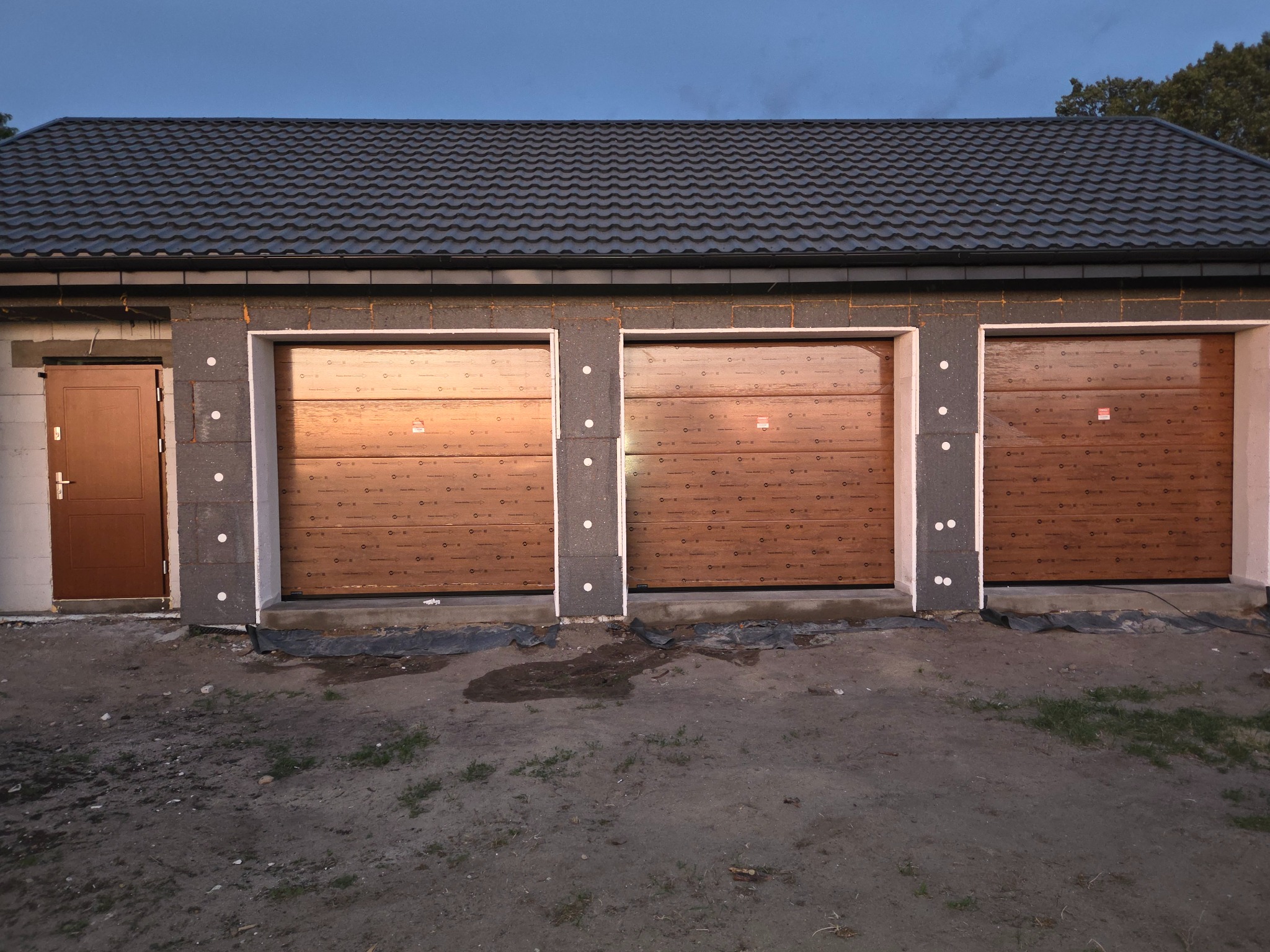 Trzy brązowe bramy garażowe segmentowe z poziomym wzorem drewna, oddzielone szarymi słupkami z izolacją, w surowym stanie wykończenia budynku.