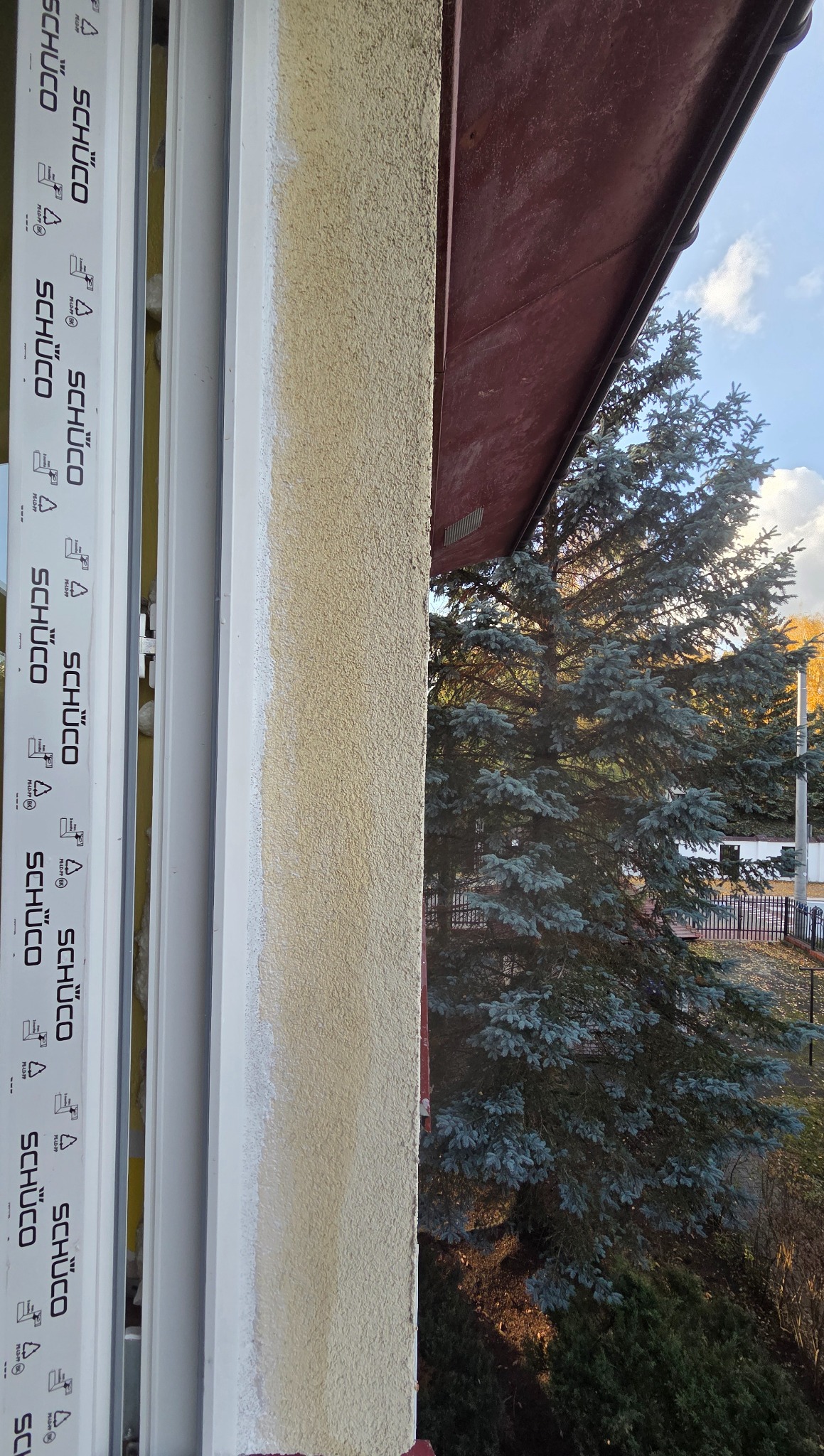Zamontowane okno PCV marki Schüco z widokiem na zewnątrz, z fragmentem elewacji o fakturze baranka i fragmentem dachu. Widoczne drzewo iglaste i błękitne niebo.