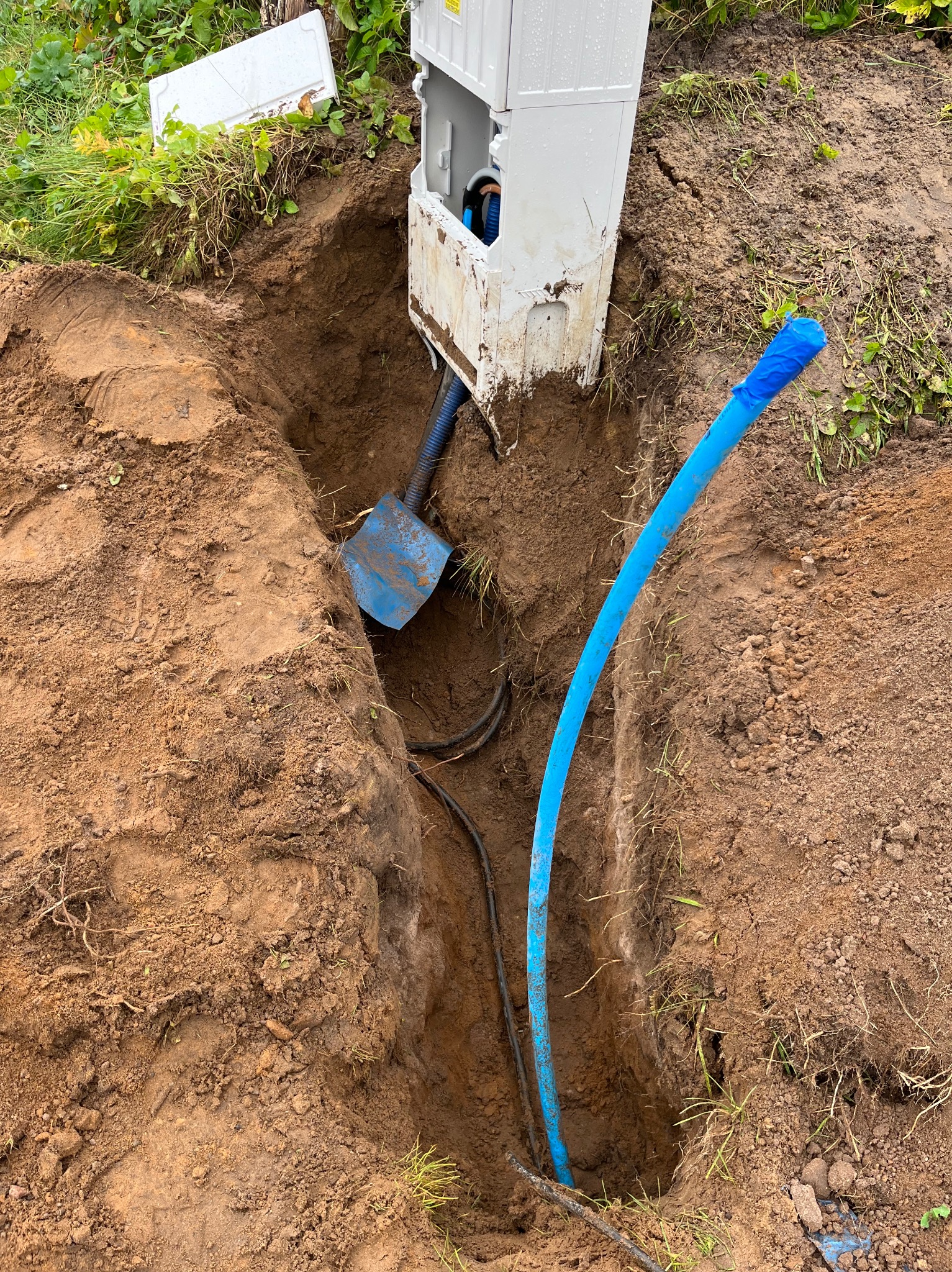 Wykop z kablem i rurą w ziemi, skrzynka elektryczna wystaje z wykopu. Widoczna łopata. Prace ziemne związane z instalacją elektryczną.
