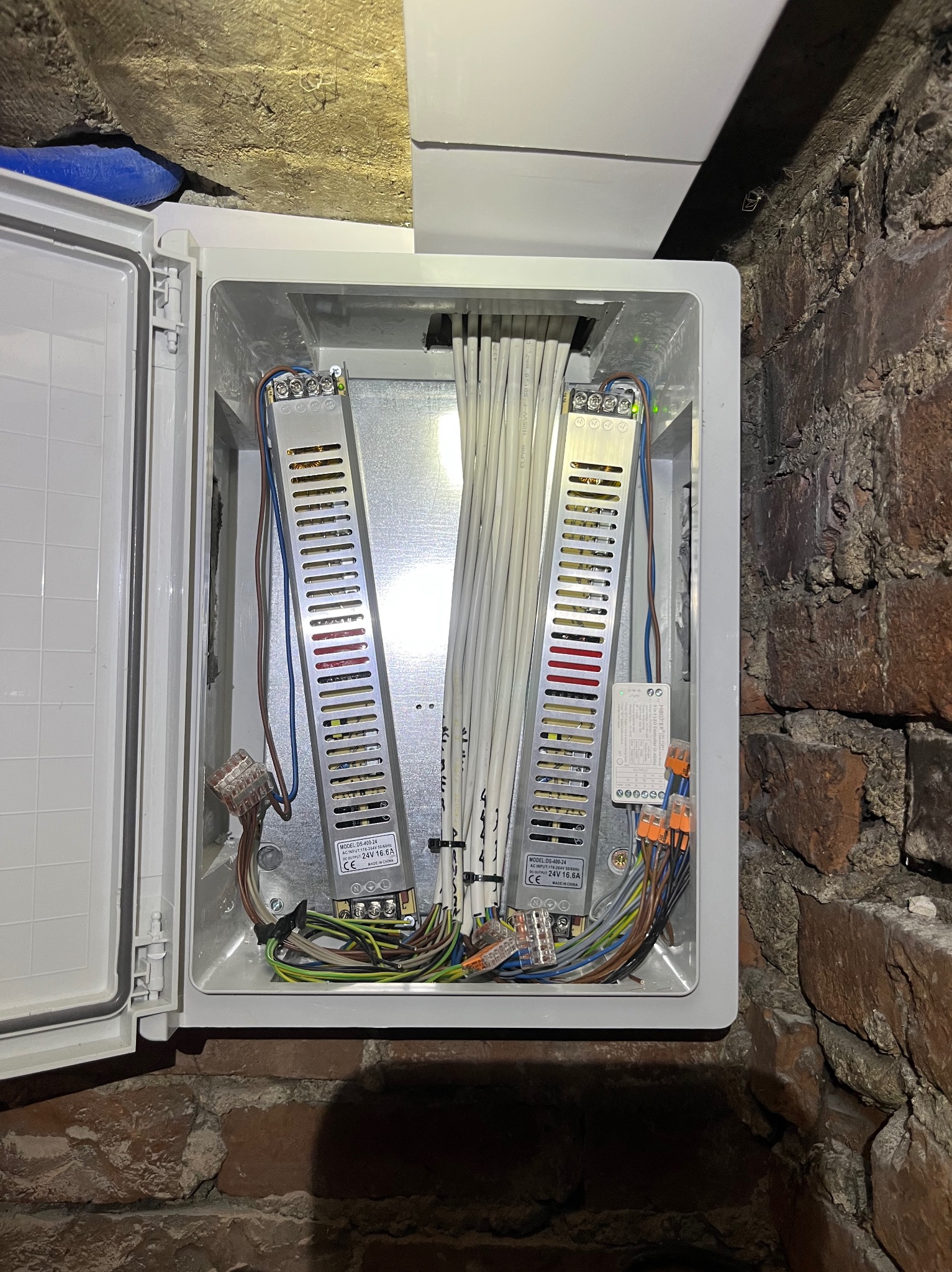 Otwarta skrzynka elektryczna z dwoma zasilaczami i wiązką kabli na tle ceglanej ściany i sufitu. Widoczne złącza WAGO i oznaczenia na zasilaczach.