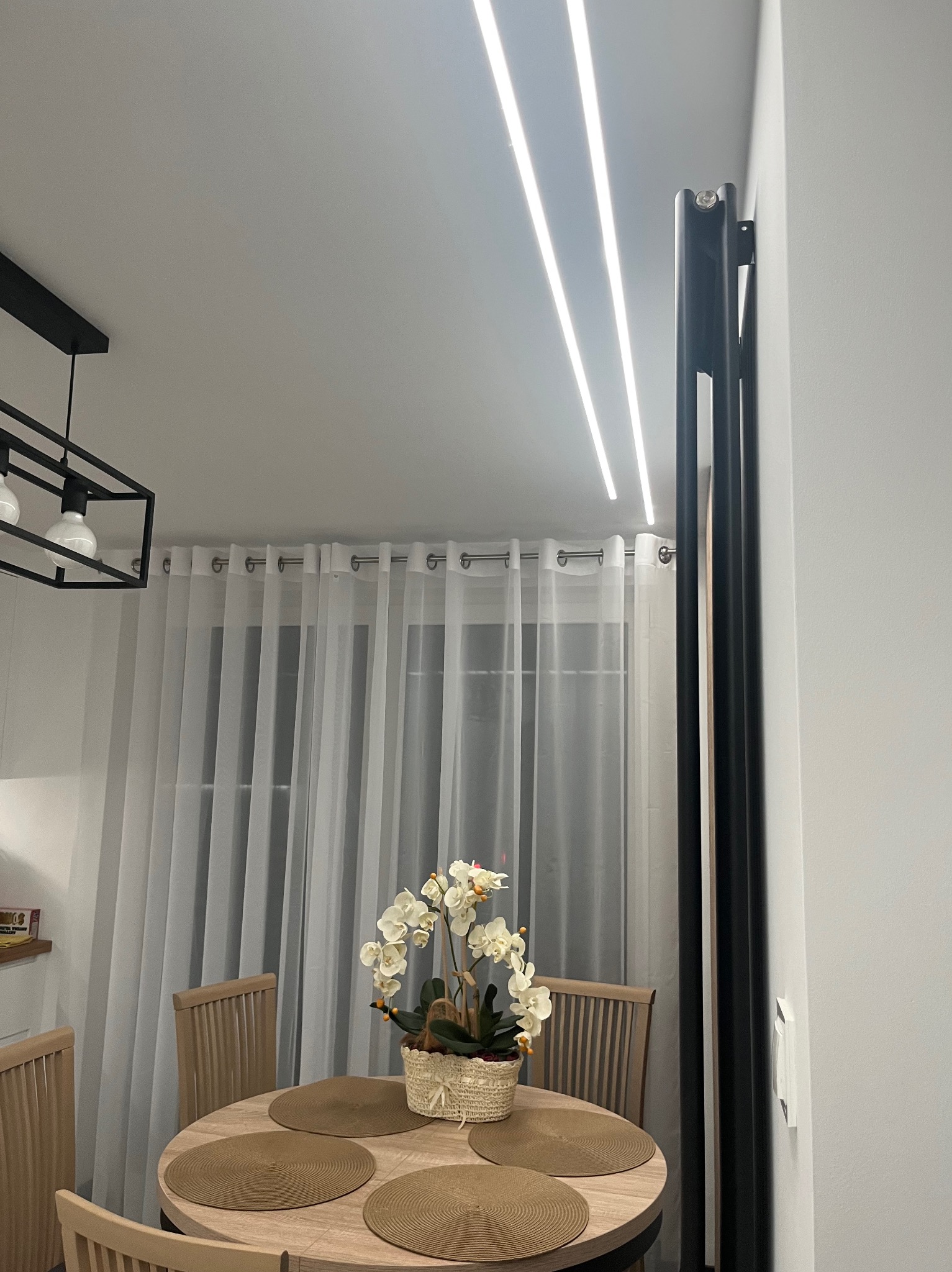 Nowoczesne oświetlenie sufitowe LED w jadalni z okrągłym stołem i kwiatami. Minimalistyczny design z białymi zasłonami w tle i czarną lampą wiszącą.