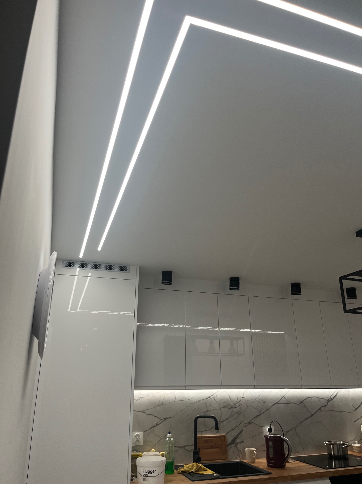 Nowoczesna kuchnia z białymi szafkami i marmurowym blatem, oświetlona liniowymi lampami LED na suficie i punktowymi reflektorami. Minimalistyczny design.