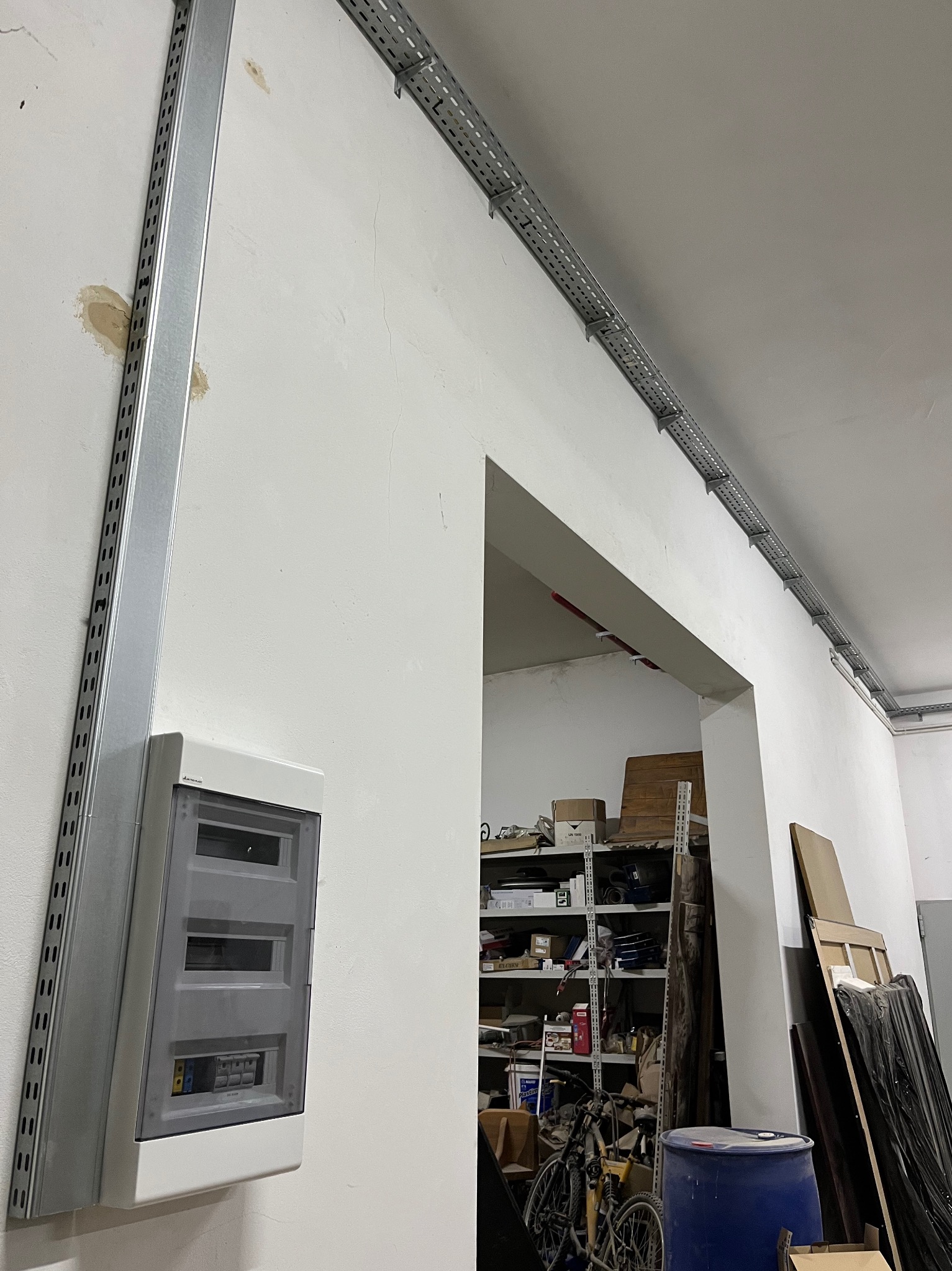 Szara skrzynka elektryczna z listwą montażową na ścianie, widoczne koryta kablowe pod sufitem. W tle zagracone pomieszczenie.