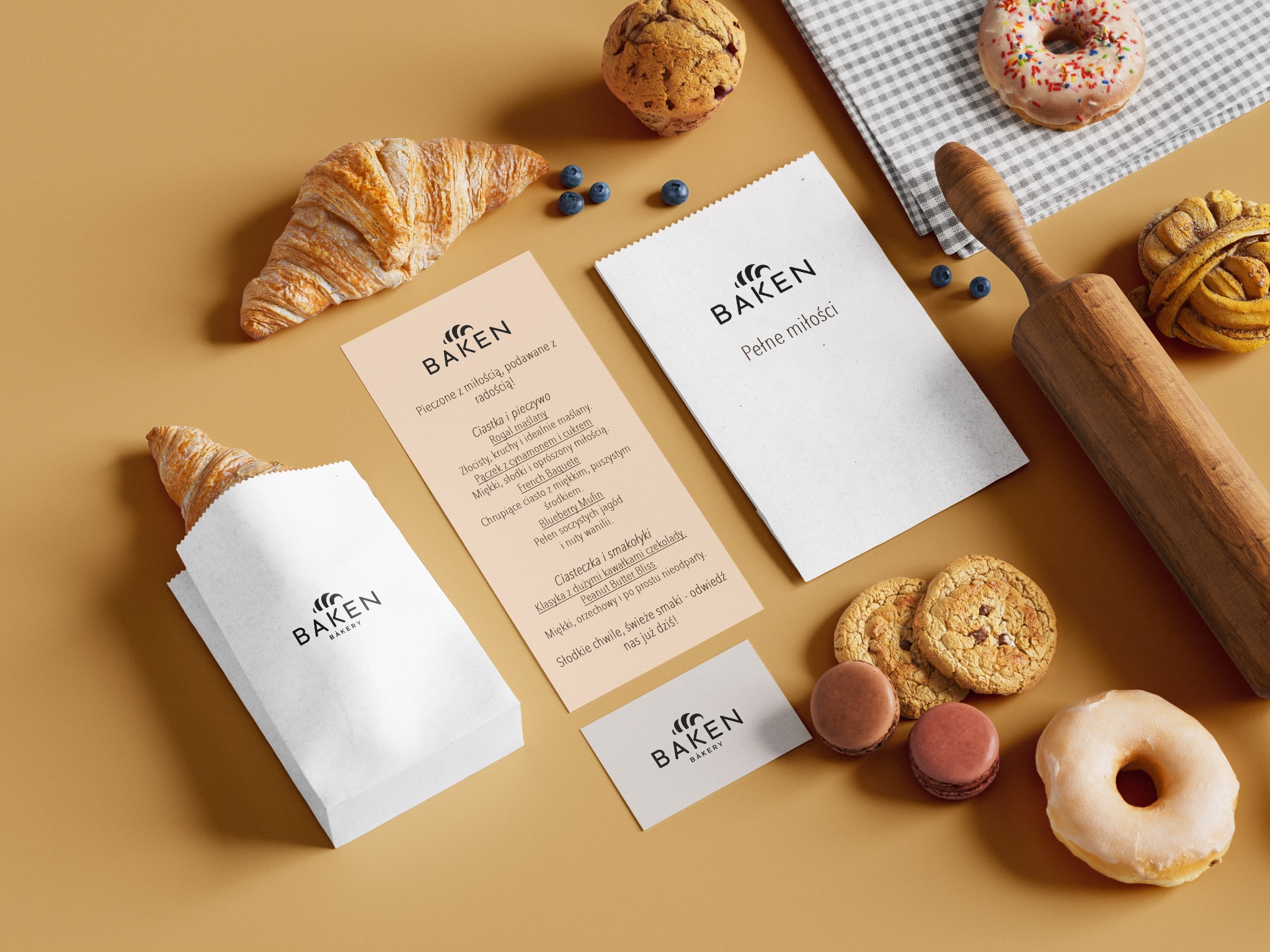 Flat lay z logo piekarni Baken na torbie, wizytówce i ulotce menu, otoczone rogalikiem, muffinką, pączkami, makaronikami i wałkiem na beżowym tle.