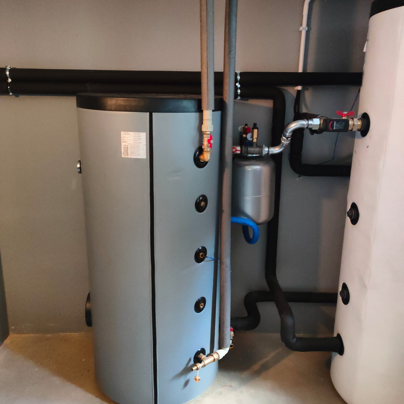 Szary bojler z podłączeniami rur i zaworami w kotłowni. Widoczne czarne rury izolowane i biały zbiornik. Instalacja grzewcza w szarym pomieszczeniu.