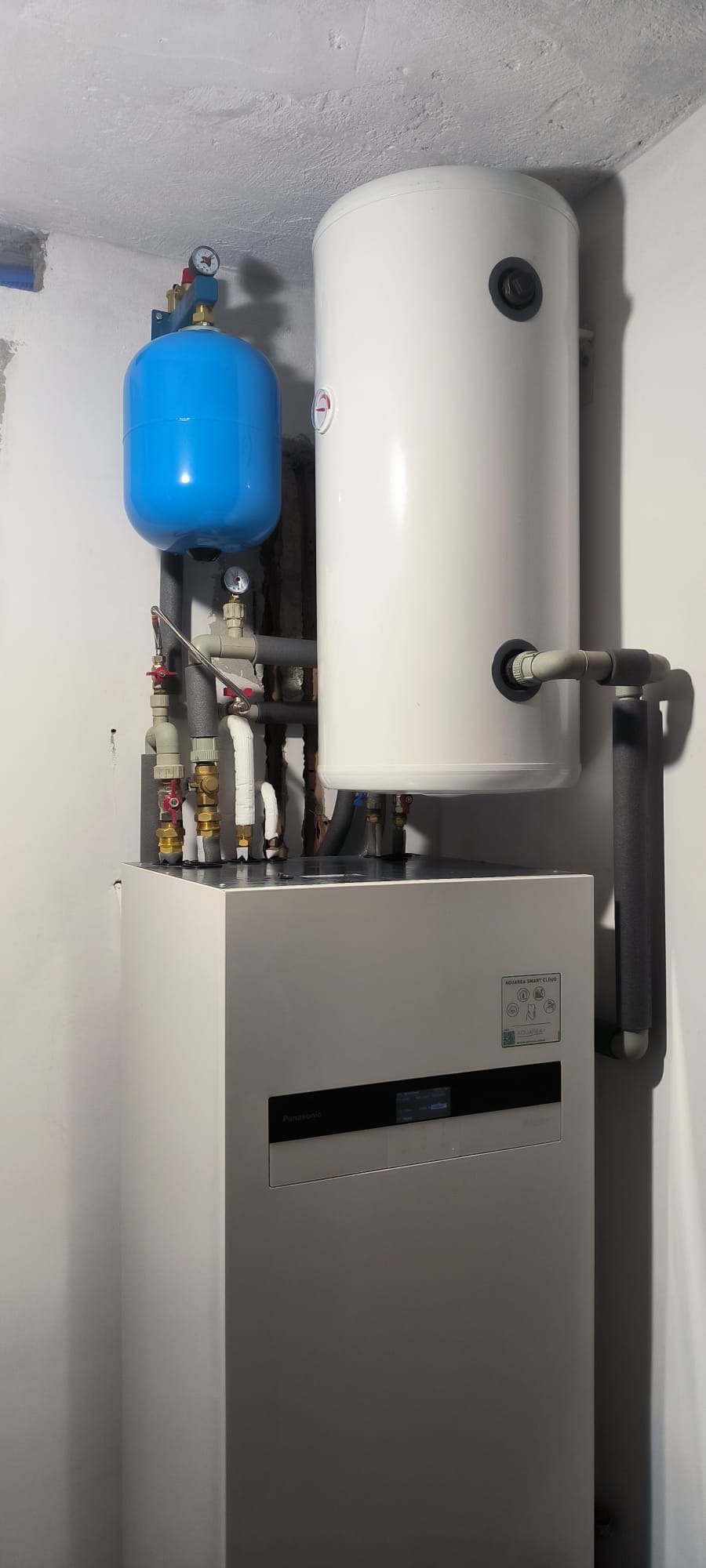 Biała pompa ciepła Panasonic Aquarea Smart Cloud z niebieskim zbiornikiem wyrównawczym i białym zasobnikiem ciepłej wody w rogu pomieszczenia.