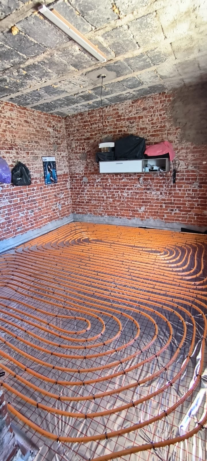 Pomarańczowe rury ogrzewania podłogowego ułożone na folii, widoczne na tle ceglanej ściany i surowego sufitu. Instalacja w trakcie realizacji.