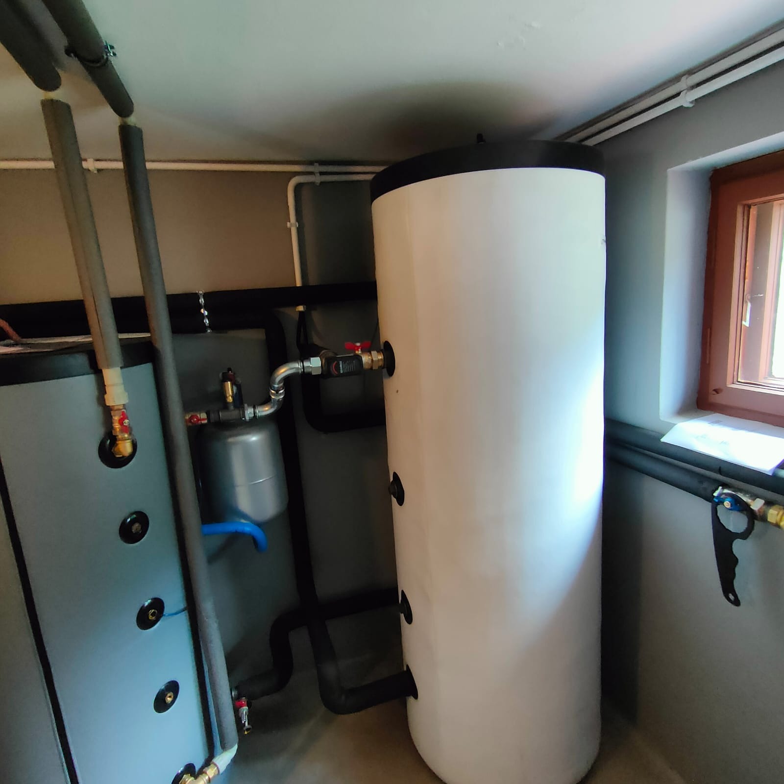 Biały zasobnik ciepłej wody użytkowej z czarnym wykończeniem, połączony z szarą instalacją grzewczą w kotłowni z oknem. Widoczne rury i zawory.