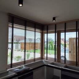 ROLFIX Jordan Frank - Drewniane żaluzje poziome w jasnej kuchni, widok na zewnątrz przez okno. Częściowo zamontowane szafki kuchenne. Dwa czarne reflektory sufitowe.