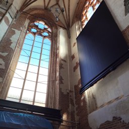 ROLFIX Jordan Frank - Wysokie okno gotyckie z zamontowaną, czarną roletą wewnętrzną. Widok z dołu na ceglane ściany i sklepienie kościoła. Część okna zasłonięta rusztowaniem.