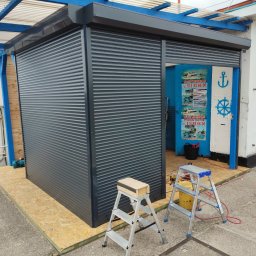 ROLFIX Jordan Frank - Szara roleta zewnętrzna zamontowana przy wejściu do budynku portowego. Widoczne drabiny i narzędzia sugerują trwający montaż. Charakterystyczne niebieskie akcenty.