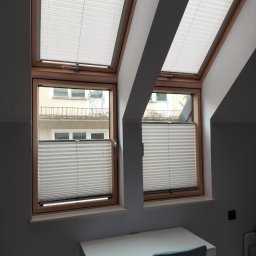 ROLFIX Jordan Frank - Dwa okna dachowe z zamontowanymi białymi roletami plisowanymi, częściowo zasłoniętymi. Widok na budynek z zewnątrz. Białe biurko i krzesło w pokoju na poddaszu.