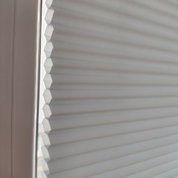 ROLFIX Jordan Frank - Delikatne, białe rolety plisowane w oknie, widoczne z bliska, zapewniające subtelne światło we Władysławowie. Charakterystyczna faktura materiału.