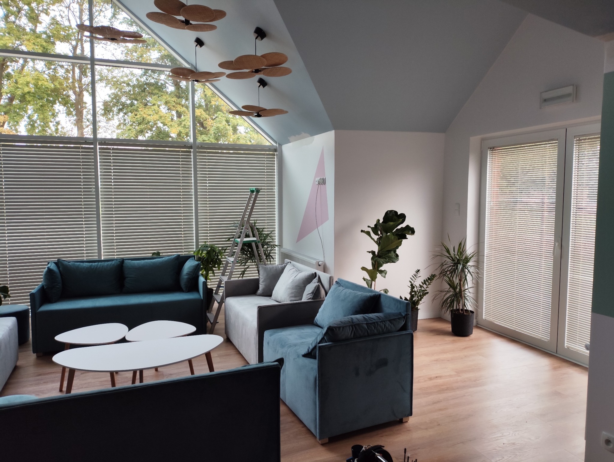 Jasne wnętrze z dużymi oknami przesłoniętymi białymi żaluzjami poziomymi. W tle drabina, sofa i rośliny doniczkowe. Widoczne elementy dekoracyjne na suficie.