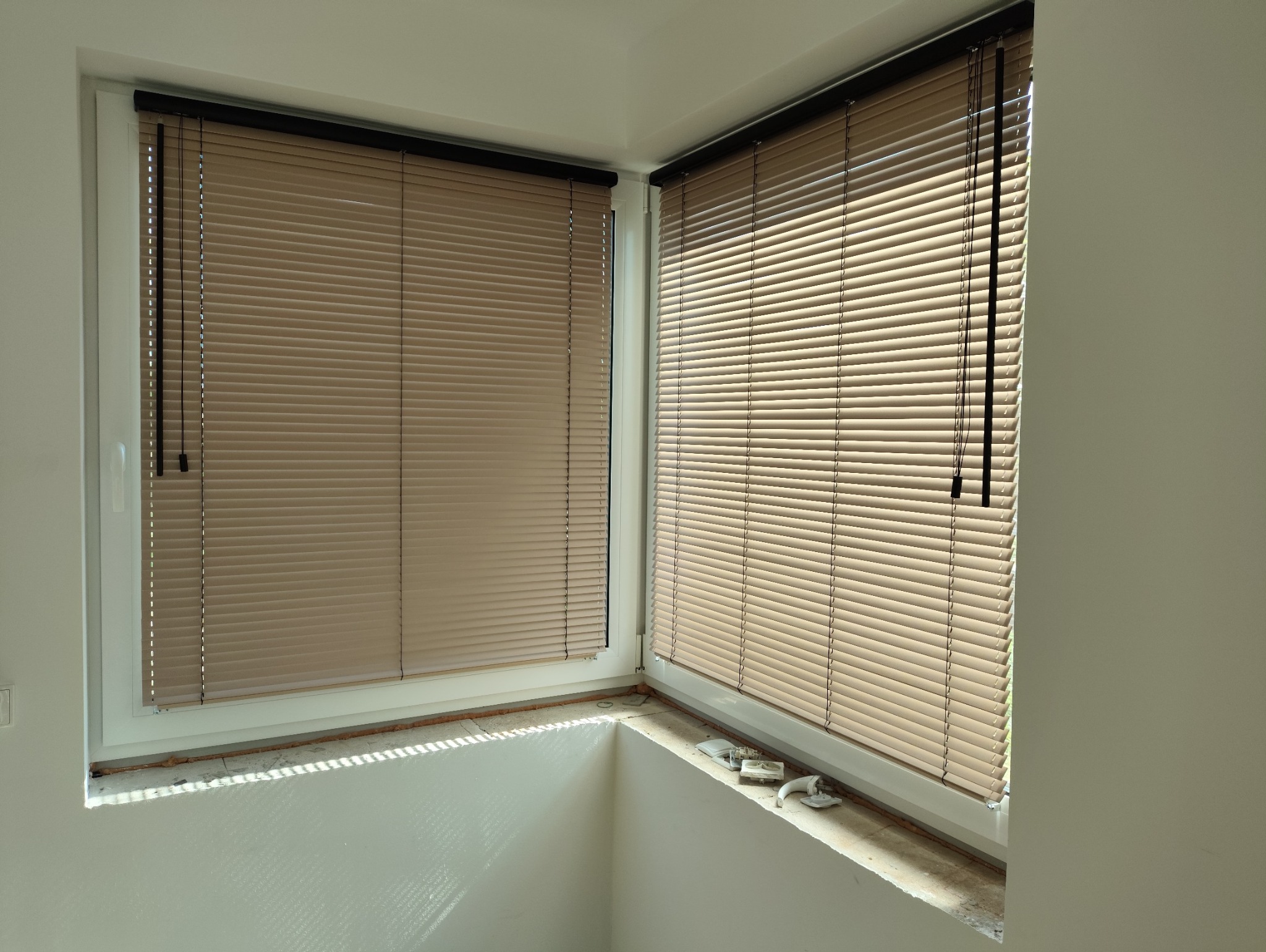 Beżowe żaluzje aluminiowe zamontowane w narożnym oknie, widoczne sznurki sterujące. Ujęcie z dołu, podkreślające kąt okna i fakturę żaluzji.