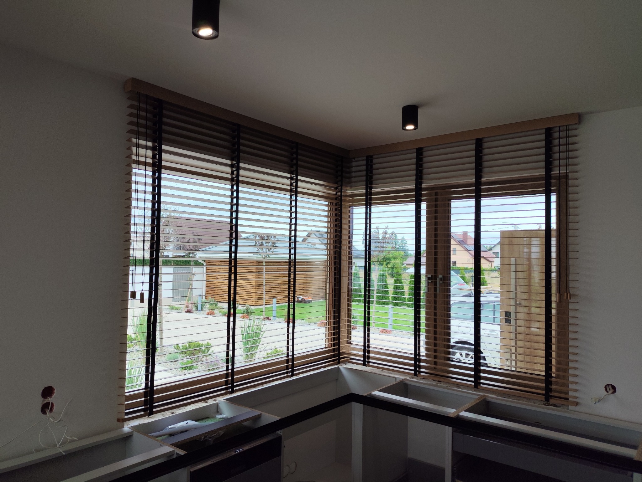 Drewniane żaluzje poziome w jasnej kuchni, widok na zewnątrz przez okno. Częściowo zamontowane szafki kuchenne. Dwa czarne reflektory sufitowe.