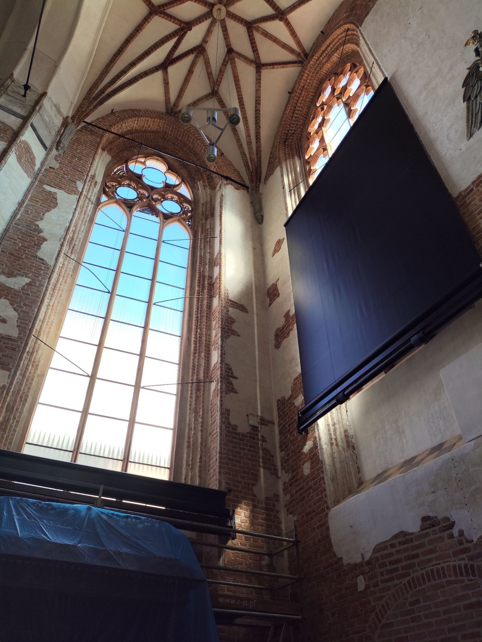 Wysokie okno gotyckie z zamontowaną, czarną roletą wewnętrzną. Widok z dołu na ceglane ściany i sklepienie kościoła. Część okna zasłonięta rusztowaniem.