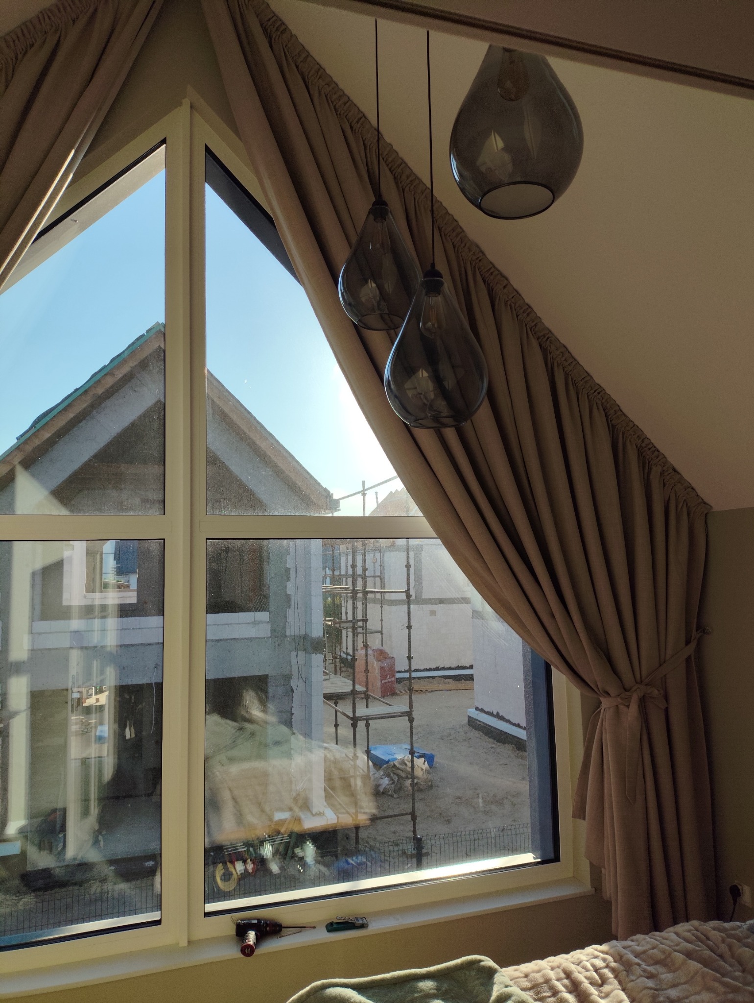 Okno z beżowymi zasłonami w pokoju na poddaszu, widok na budowę domu z rusztowaniami. Na parapecie wiertarka i nóż. Trzy szklane lampy wiszące.