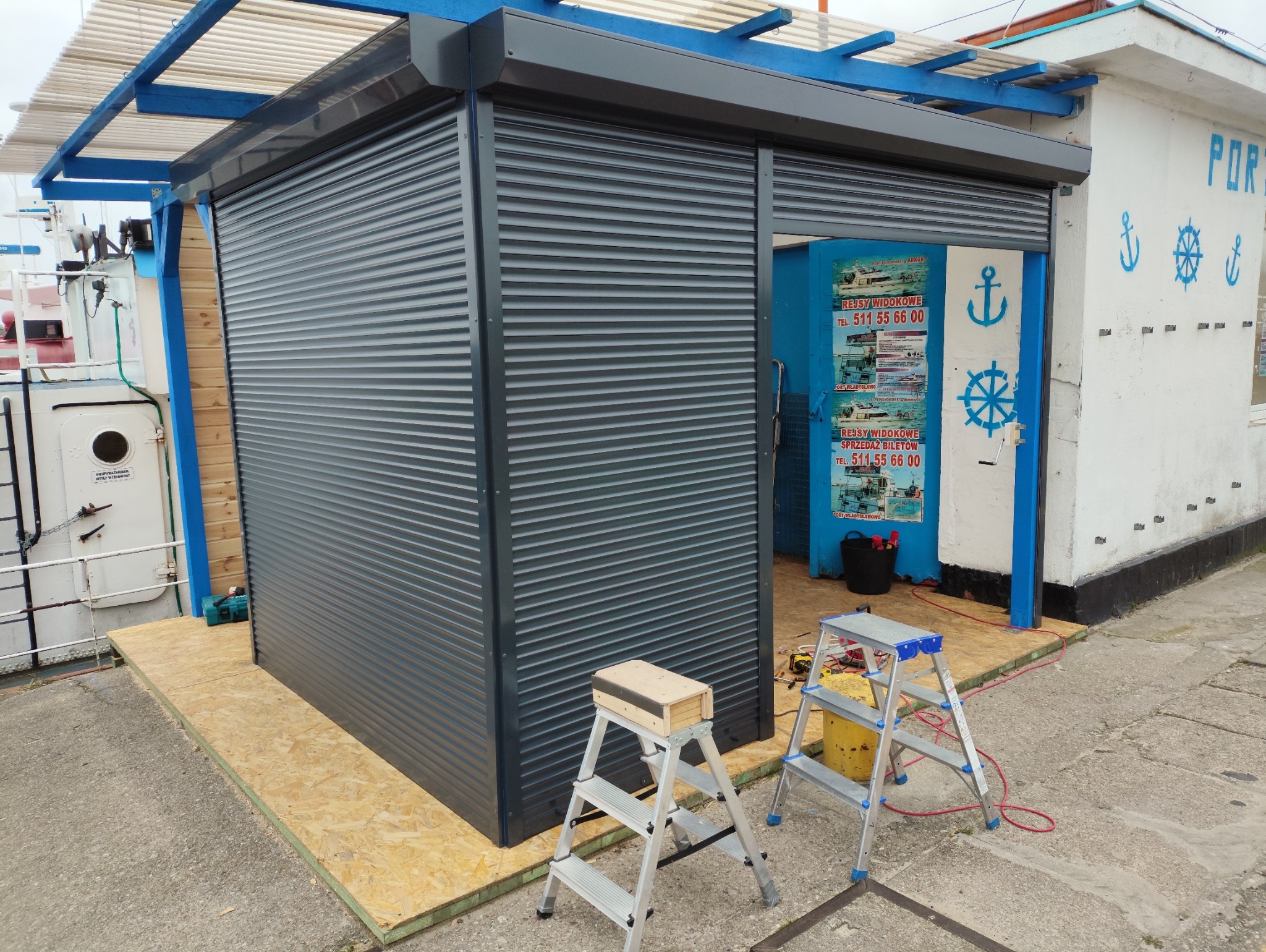 Szara roleta zewnętrzna zamontowana przy wejściu do budynku portowego. Widoczne drabiny i narzędzia sugerują trwający montaż. Charakterystyczne niebieskie akcenty.