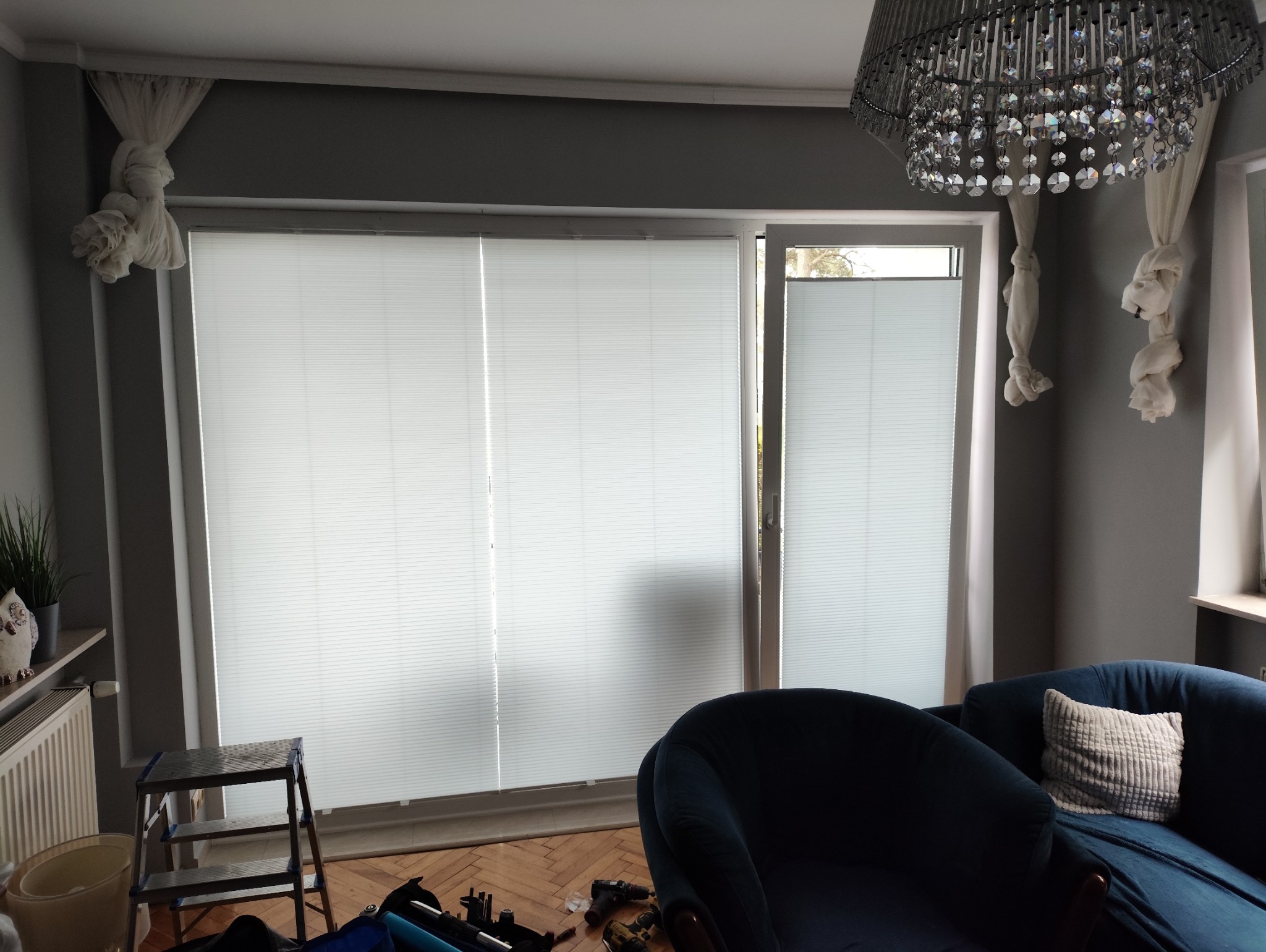 Jasne, plisowane rolety wewnętrzne zamontowane w oknie salonu. Widoczny fragment niebieskiego fotela i drabina. Dekoracyjne upięcie zasłon.