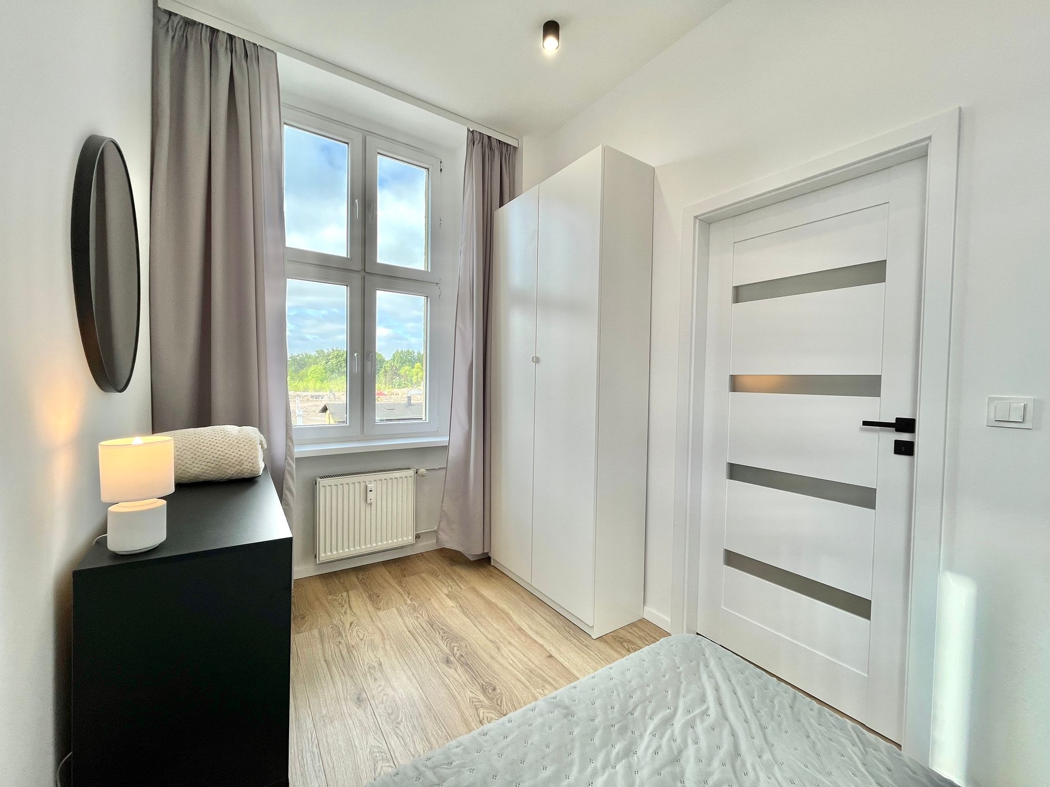 Jasne wnętrze pokoju po remoncie: białe ściany, drewniana podłoga, okno z widokiem na zieleń, minimalistyczna szafa i czarna komoda z lampką.