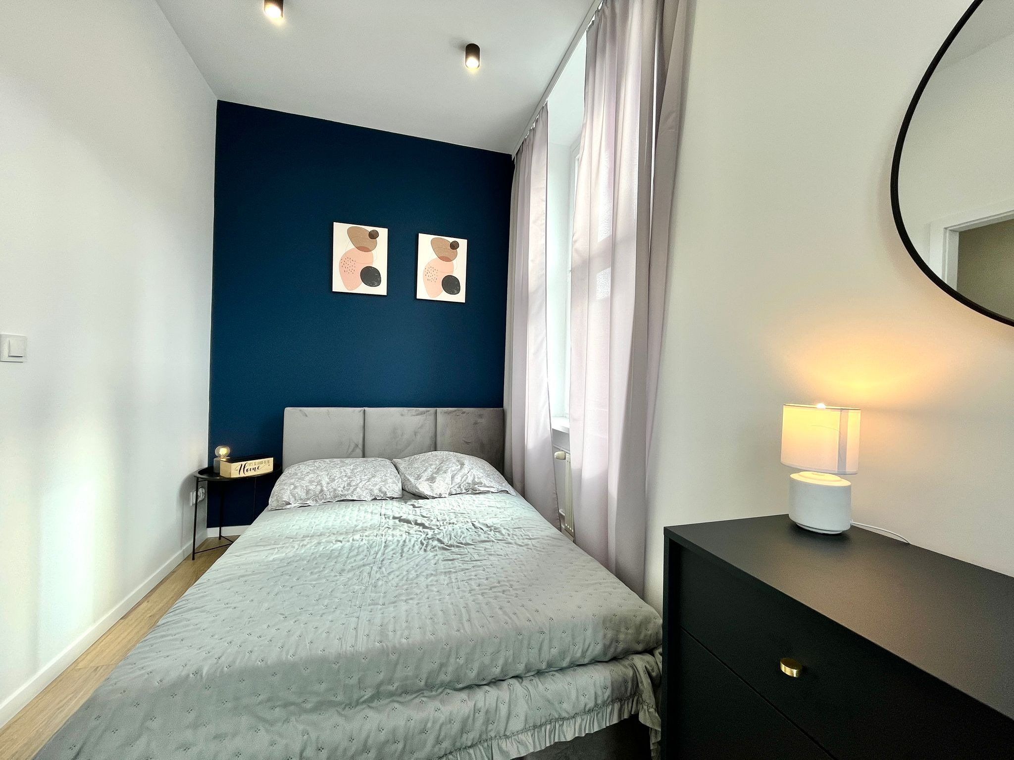 Nowoczesna sypialnia z łóżkiem, ciemnoniebieską ścianą z abstrakcyjnymi obrazami i minimalistyczną lampką nocną na czarnej komodzie. Stonowana kolorystyka i przytulny klimat.