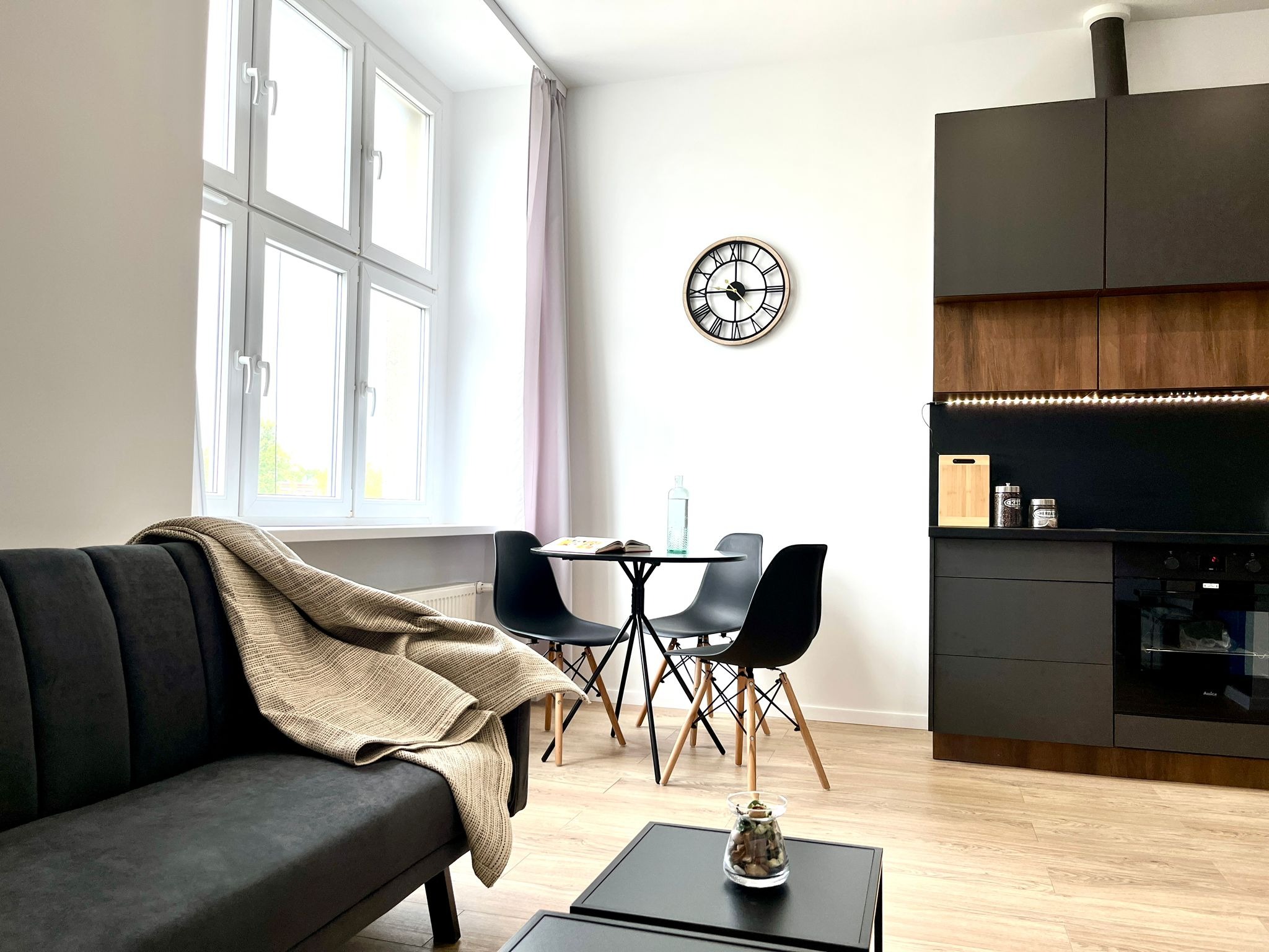 Jasne wnętrze mieszkania po remoncie: szara sofa z pledem, okrągły stół z krzesłami, czarne meble kuchenne z drewnianymi akcentami, zegar na ścianie.