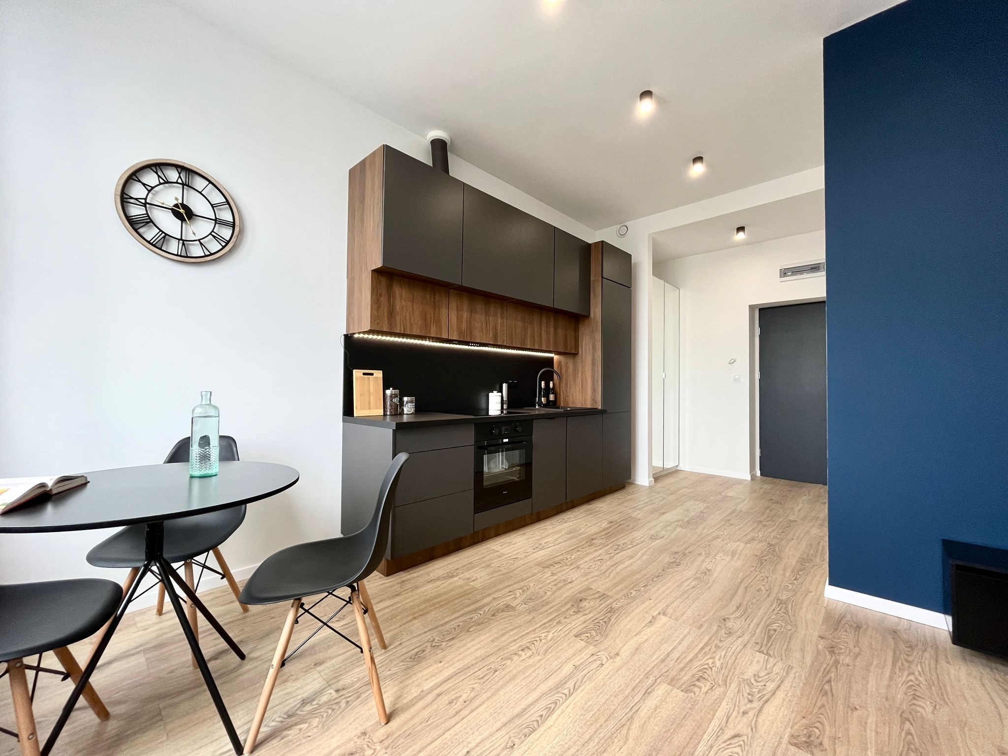 Nowoczesna kuchnia z ciemnymi szafkami i drewnianymi akcentami, okrągły stół z czarnymi krzesłami, drewniana podłoga i niebieska ściana. Minimalistyczny design.