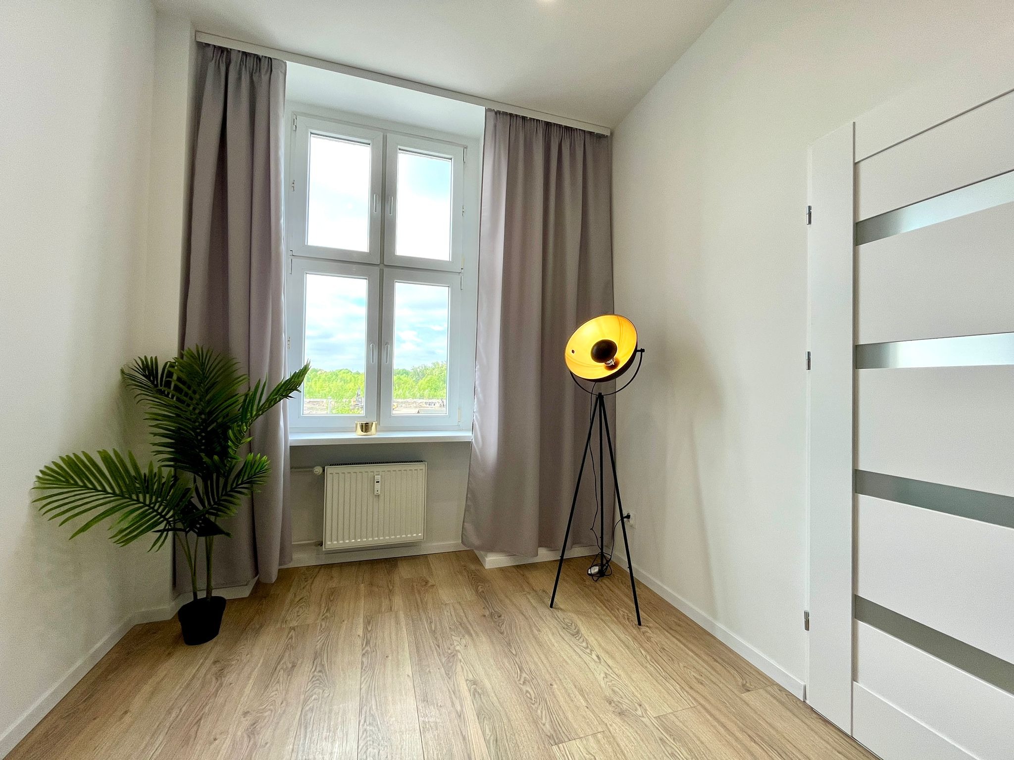 Jasne wnętrze pokoju z drewnianą podłogą, oknem z zasłonami, lampą na trójnogu i rośliną doniczkową. Minimalistyczny design z białymi ścianami i drzwiami.