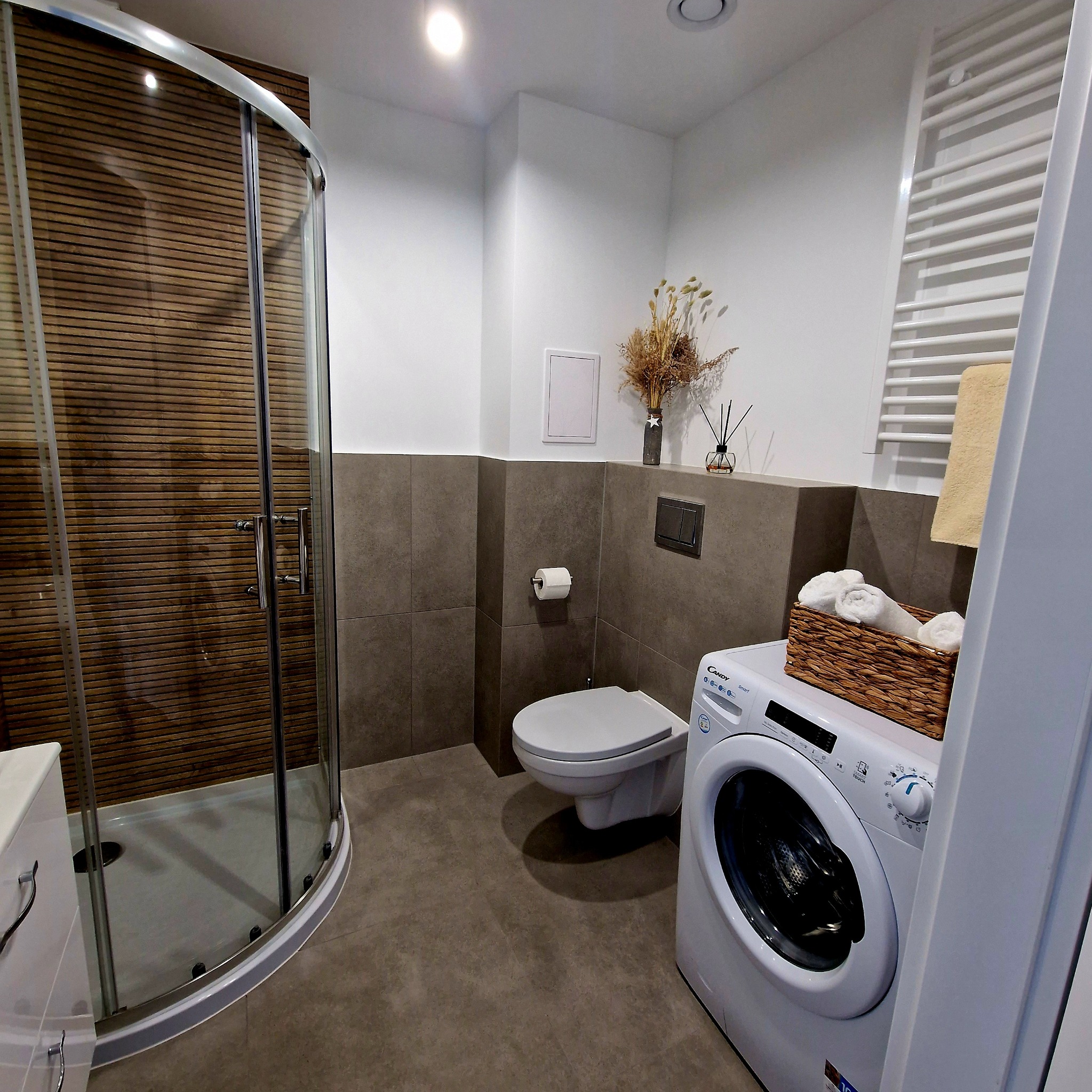 Nowoczesna łazienka z kabiną prysznicową z drewnianymi panelami, toaletą podwieszaną i pralką z koszem na ręczniki. Minimalistyczny design.