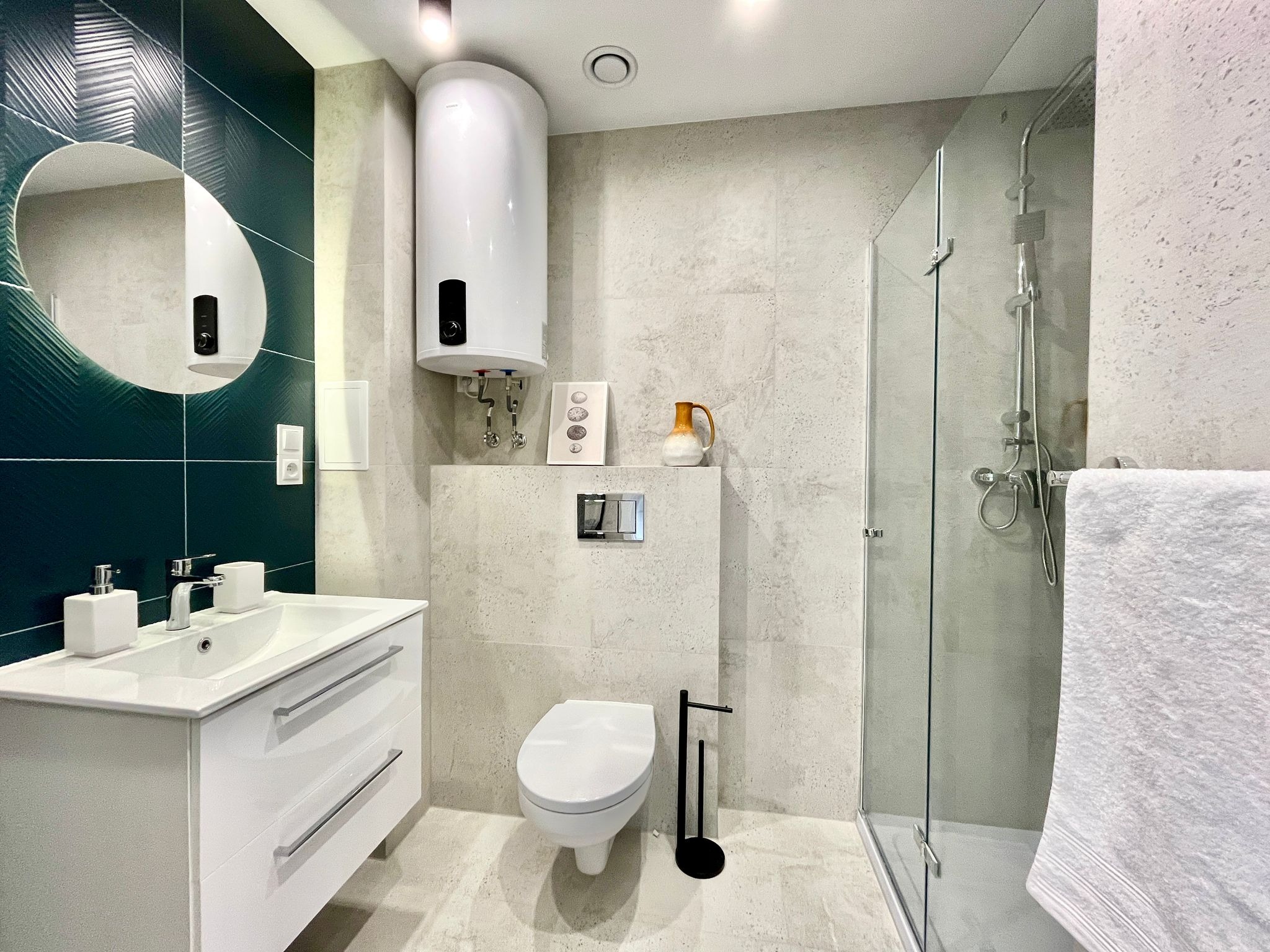 Nowoczesna łazienka z kabiną prysznicową, umywalką z szafką, toaletą i bojlerem. Ciemnozielone płytki z wzorem liści za lustrem dodają elegancji.