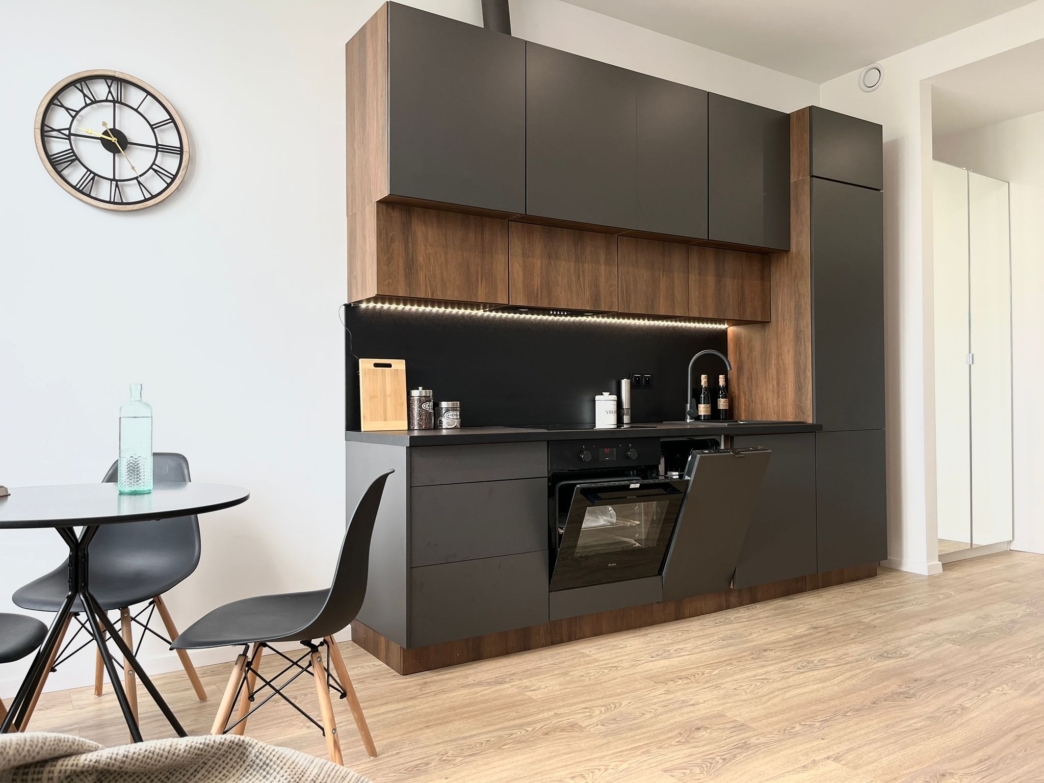 Nowoczesna kuchnia w Siemianowicach Śląskich: grafitowe szafki z drewnianymi akcentami, okrągły stół z czarnymi krzesłami, otwarty piekarnik, zegar na ścianie.
