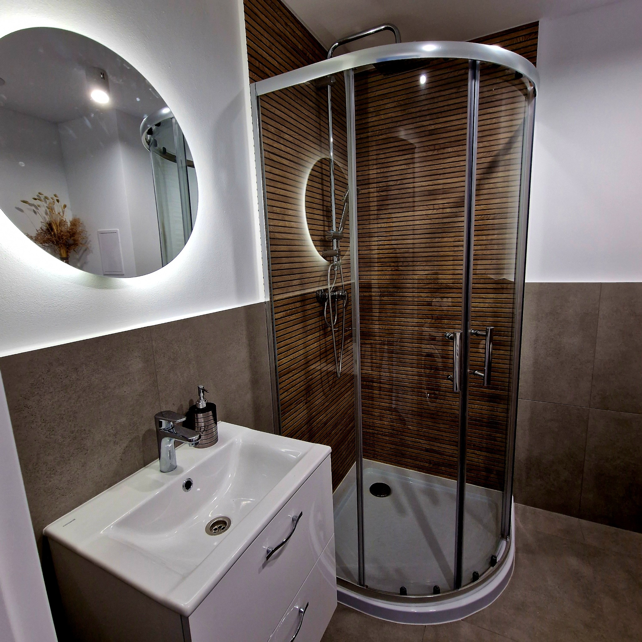 Nowoczesna łazienka z kabiną prysznicową z drewnianym panelem, umywalką z szafką i okrągłym lustrem z podświetleniem LED. Minimalistyczny design, brązowo-biała kolorystyka.