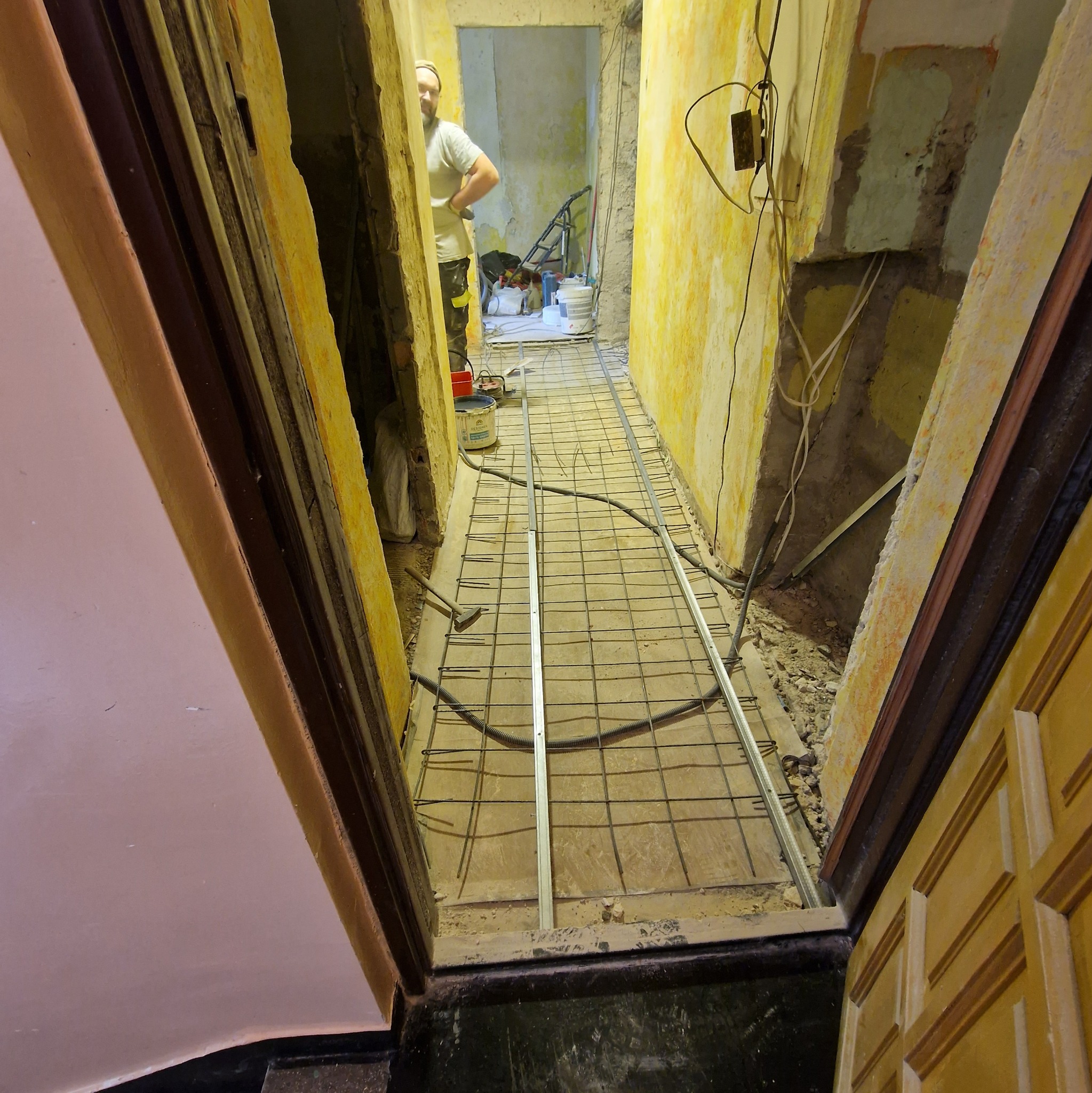 Przygotowanie podłoża pod wylewkę w korytarzu. Widoczna siatka zbrojeniowa i profile. Ściany w trakcie remontu, widoczne instalacje elektryczne. Osoba w tle.