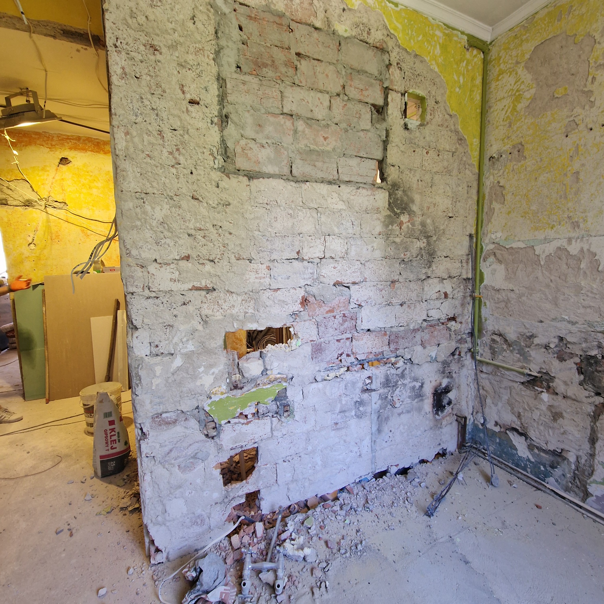 Odsłonięta ceglana ściana z ubytkami tynku w trakcie remontu. Widoczne instalacje i materiały budowlane. Żółte ściany w tle.