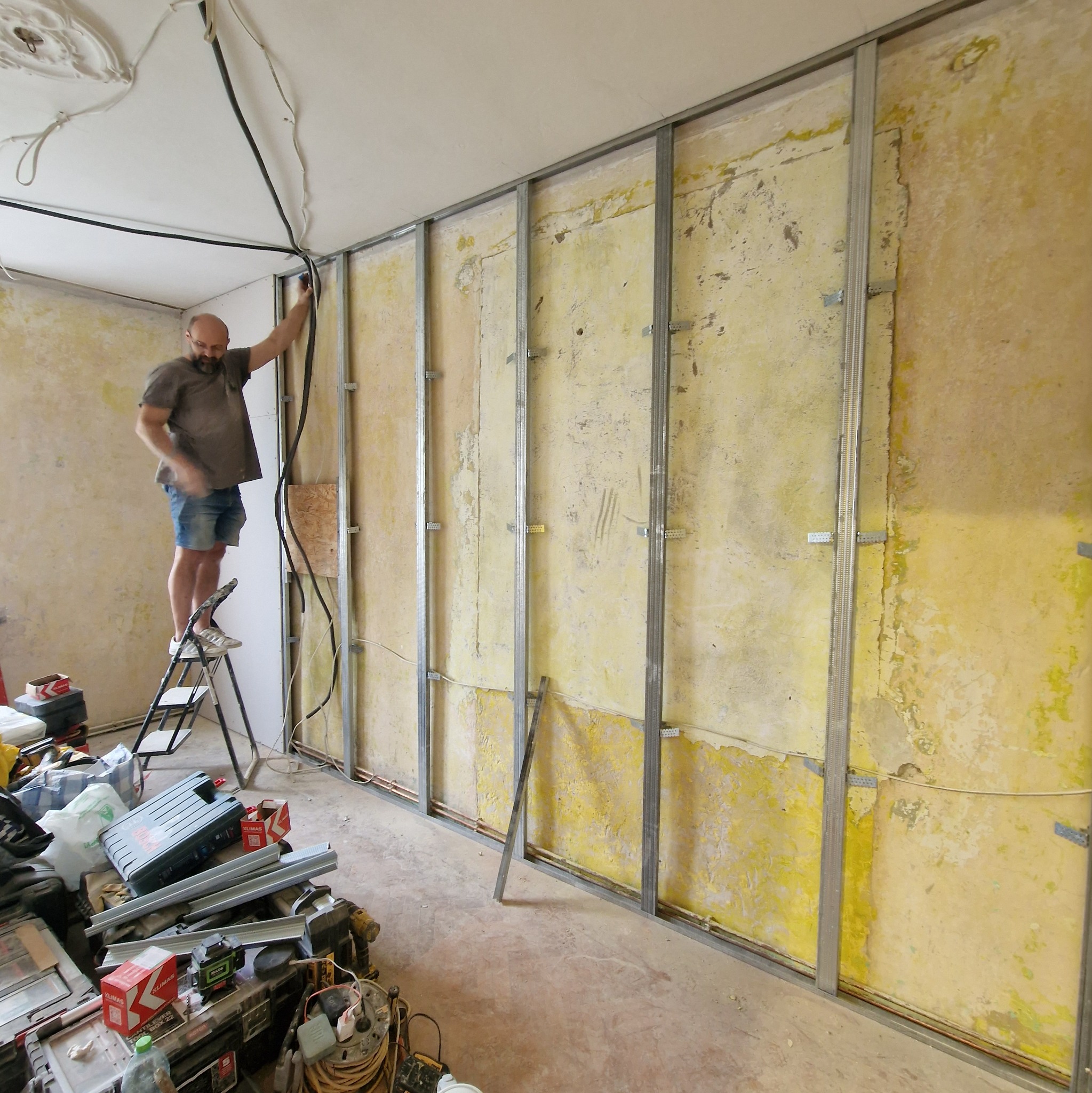 Montaż stelaża pod płytę karton-gips na starej, żółtej ścianie. Elektryk na drabinie układa kable. Na podłodze narzędzia i materiały budowlane.